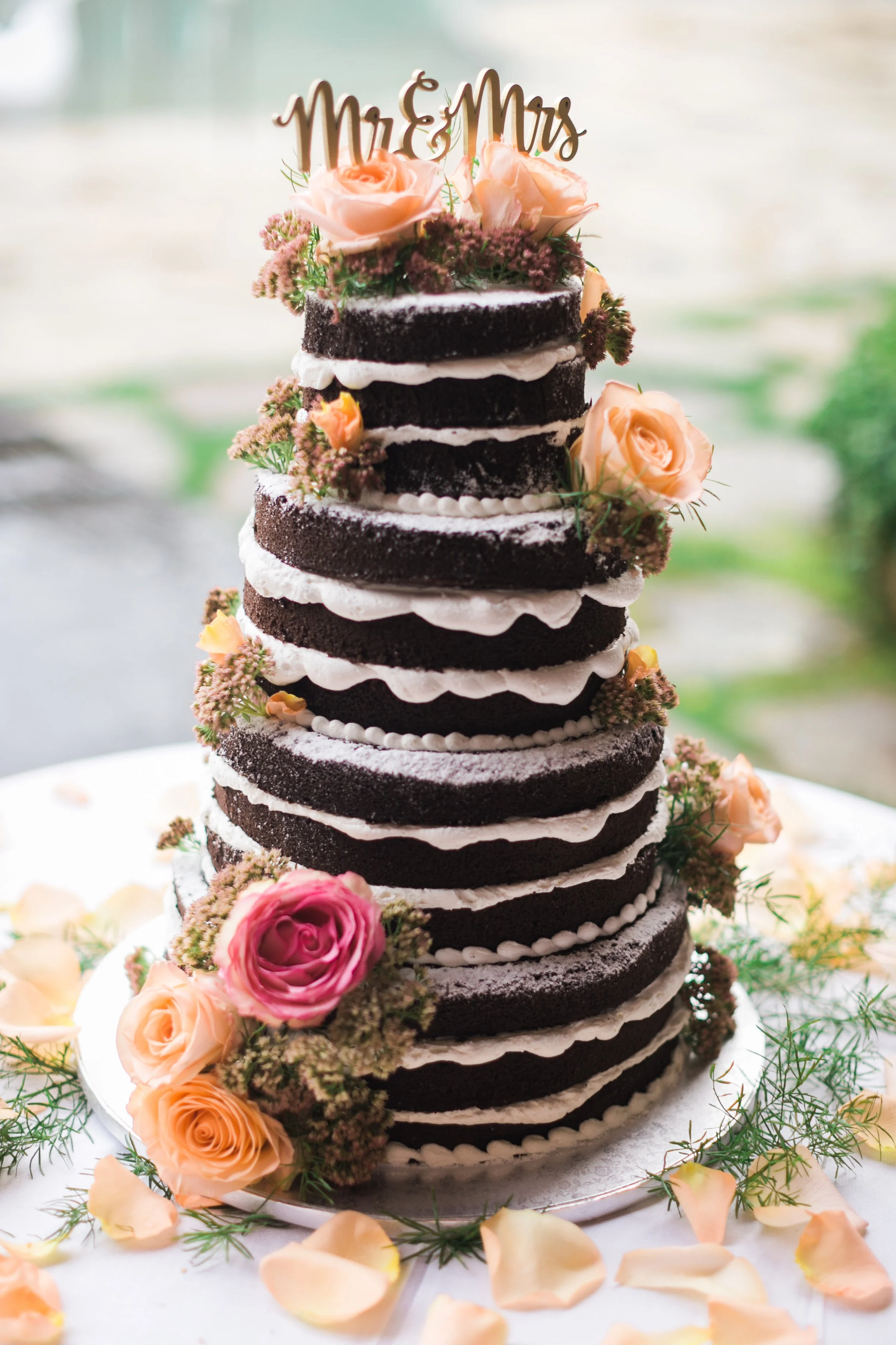 Naked wedding cake with chocolate layers, cream filling, and adorned with pink and peach roses, topped with a "Mr & Mrs" sign.