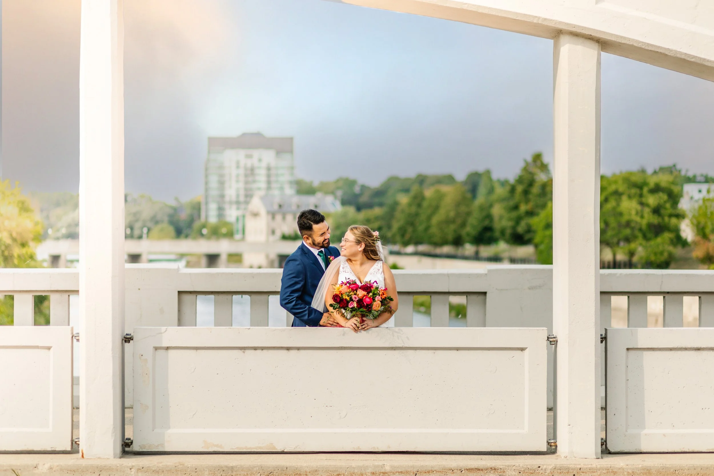 Couple in wedding attire on a bridge, holding a bouquet, smiling at each other, with a cityscape and greenery in the background.