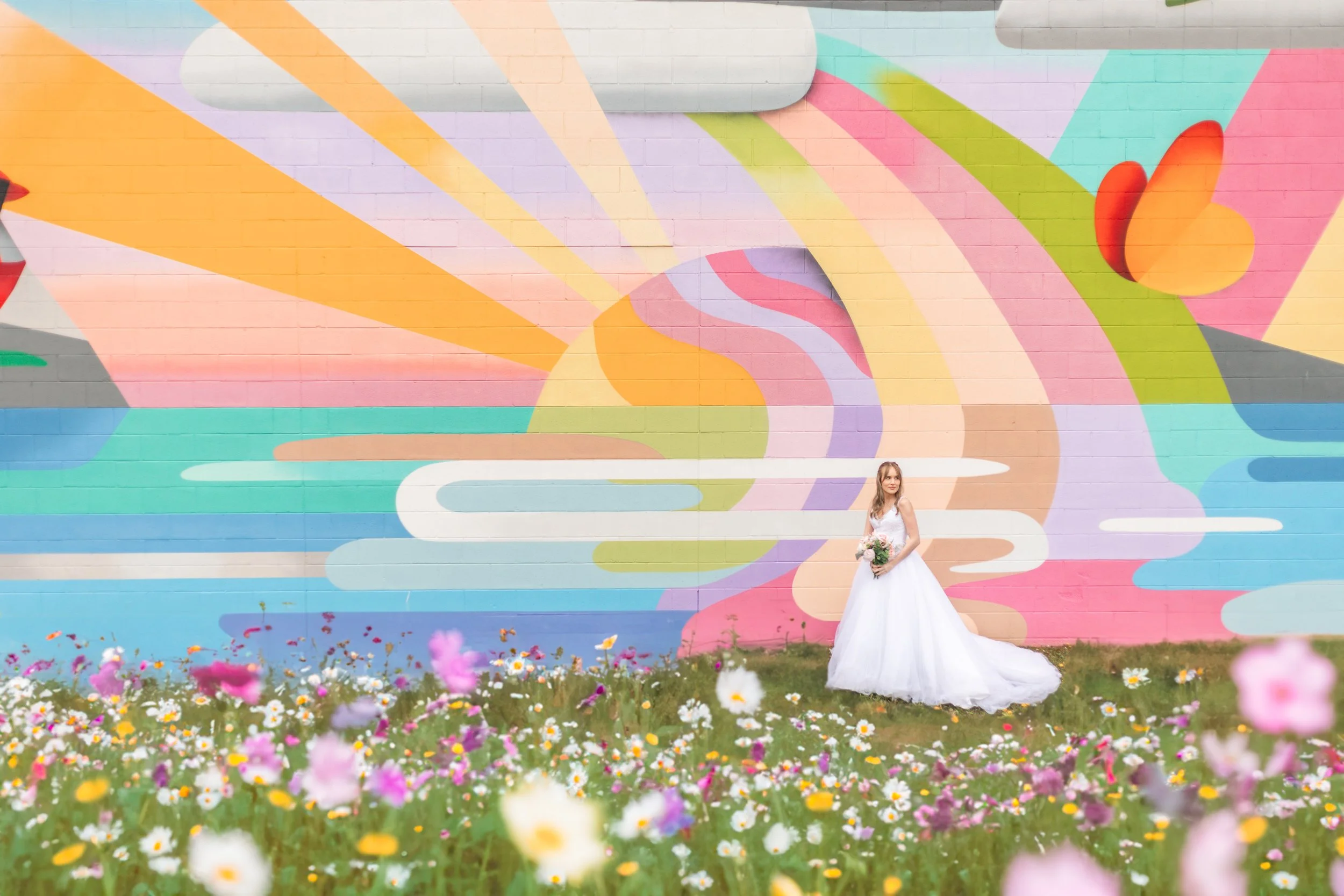 Bride standing in a field of flowers with a colorful mural backdrop featuring a sun and abstract shapes.