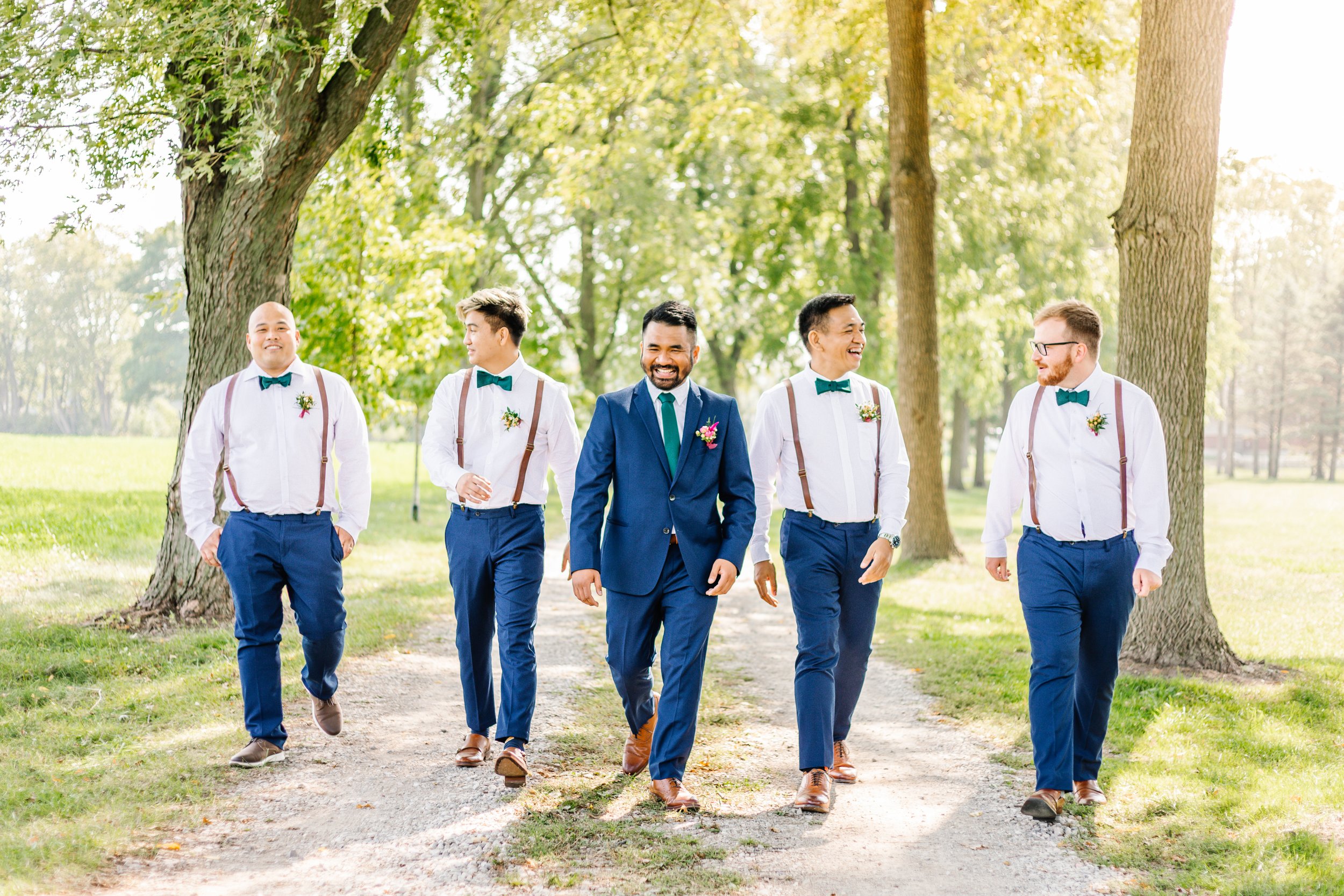 A groom in a blue suit walks with four groomsmen wearing white shirts, suspenders, and teal bow ties on a tree-lined path in a park.