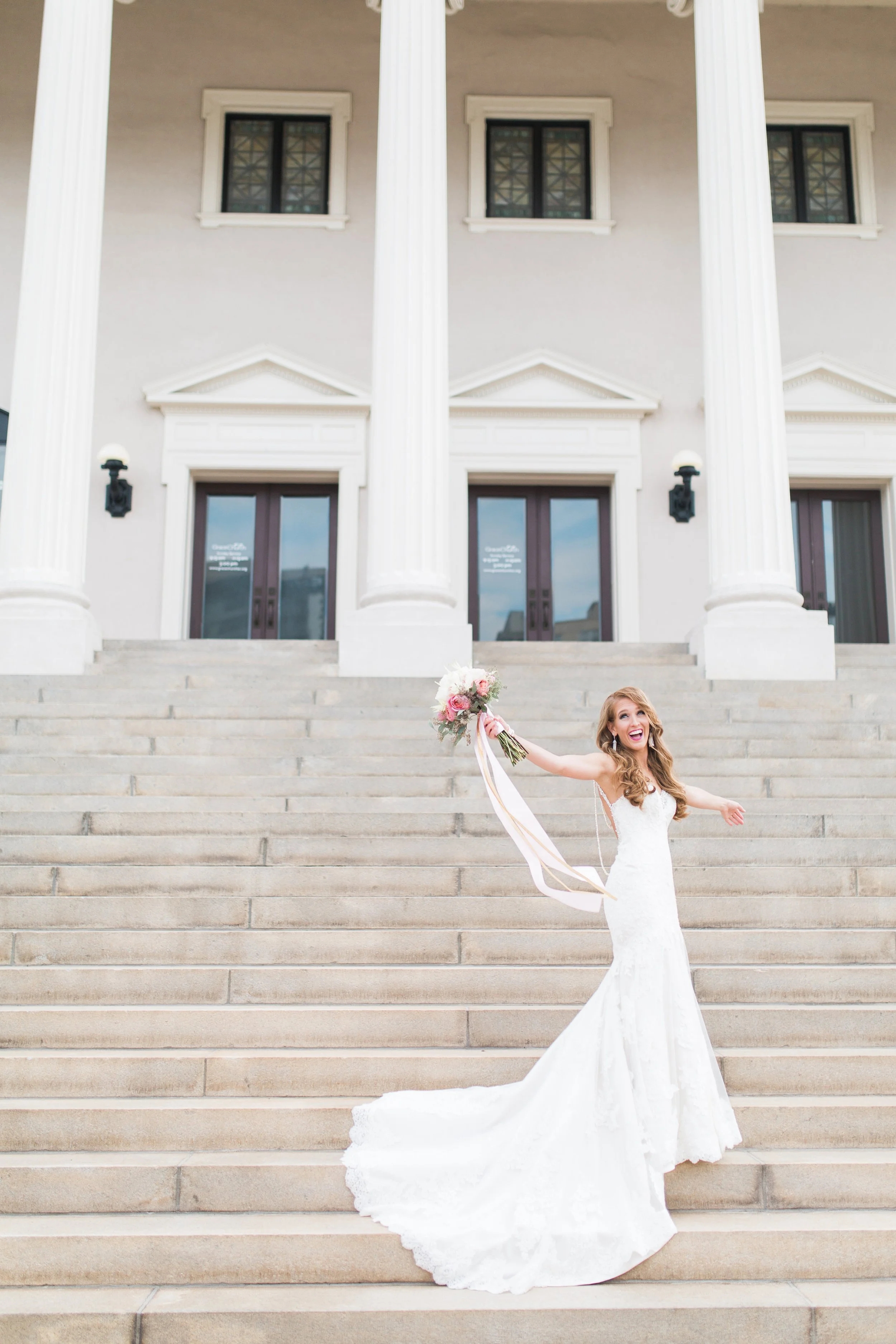 Bride in a white wedding dress holding a bouquet on stone steps in front of a building with columns.