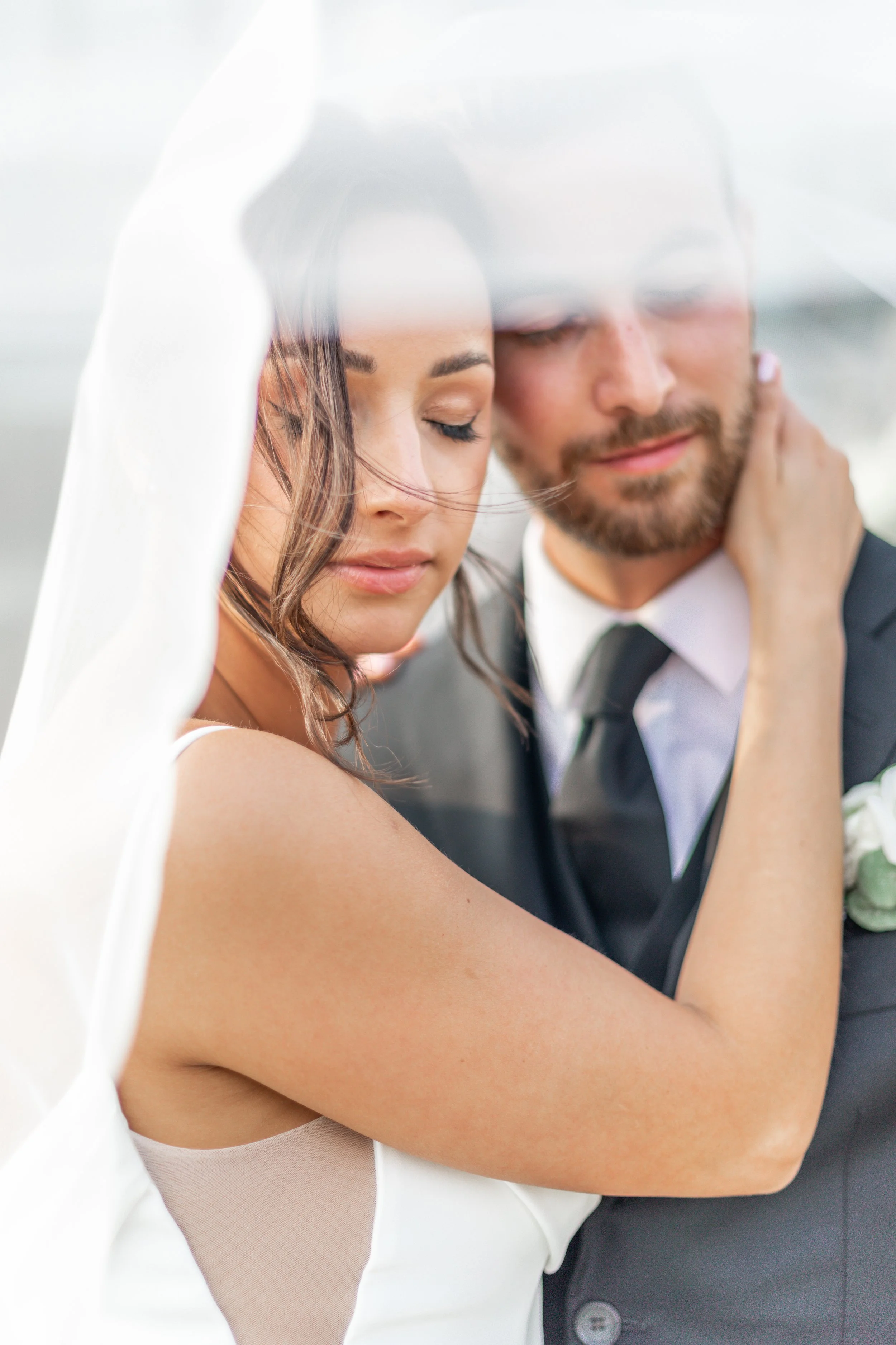 Bride and groom embracing under a veil, with eyes closed, in soft focus.