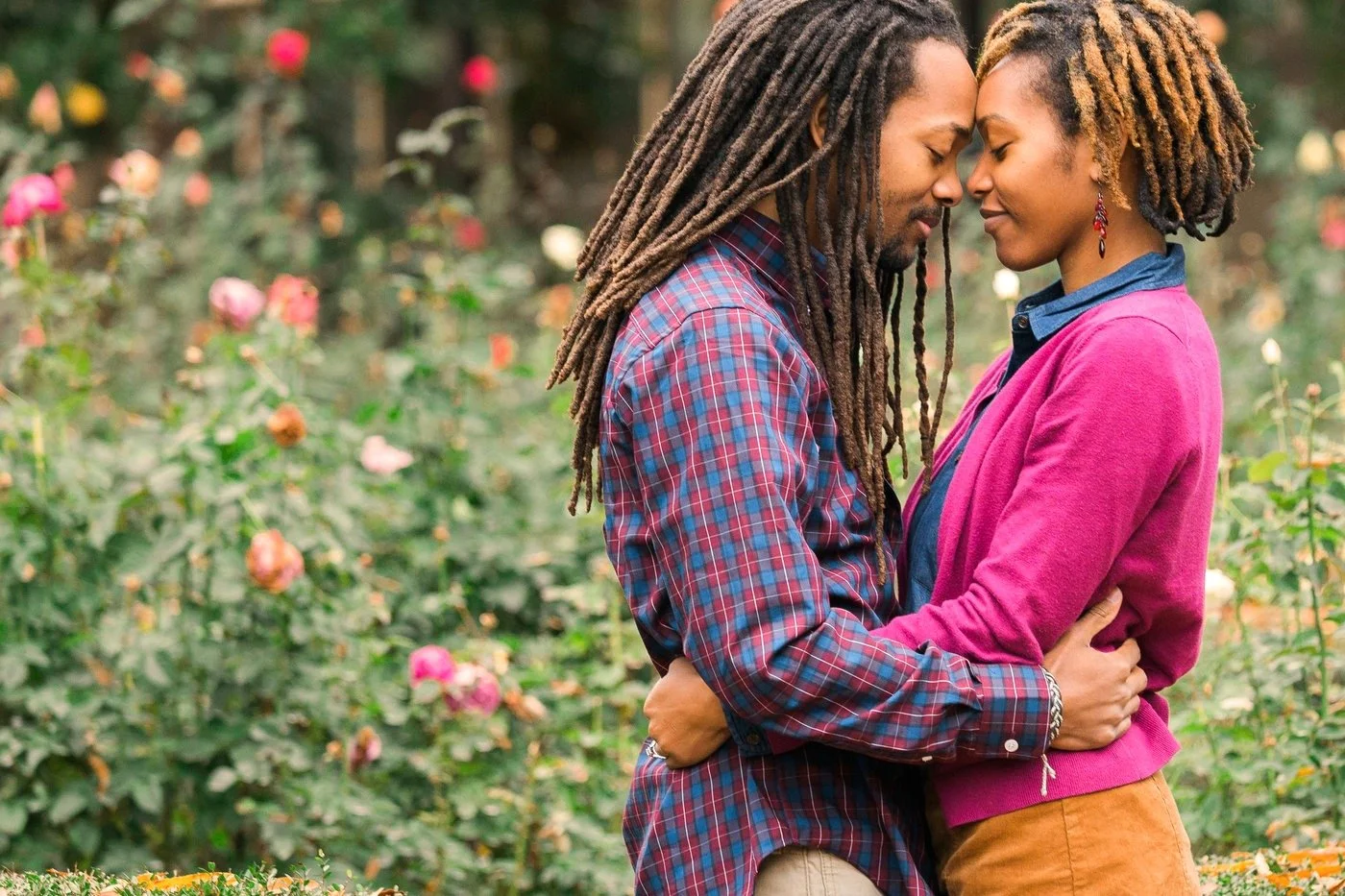 A couple embraces affectionately in a garden, with their eyes closed and foreheads touching. They are surrounded by flowers and greenery. The man wears a plaid shirt, and the woman wears a pink sweater.