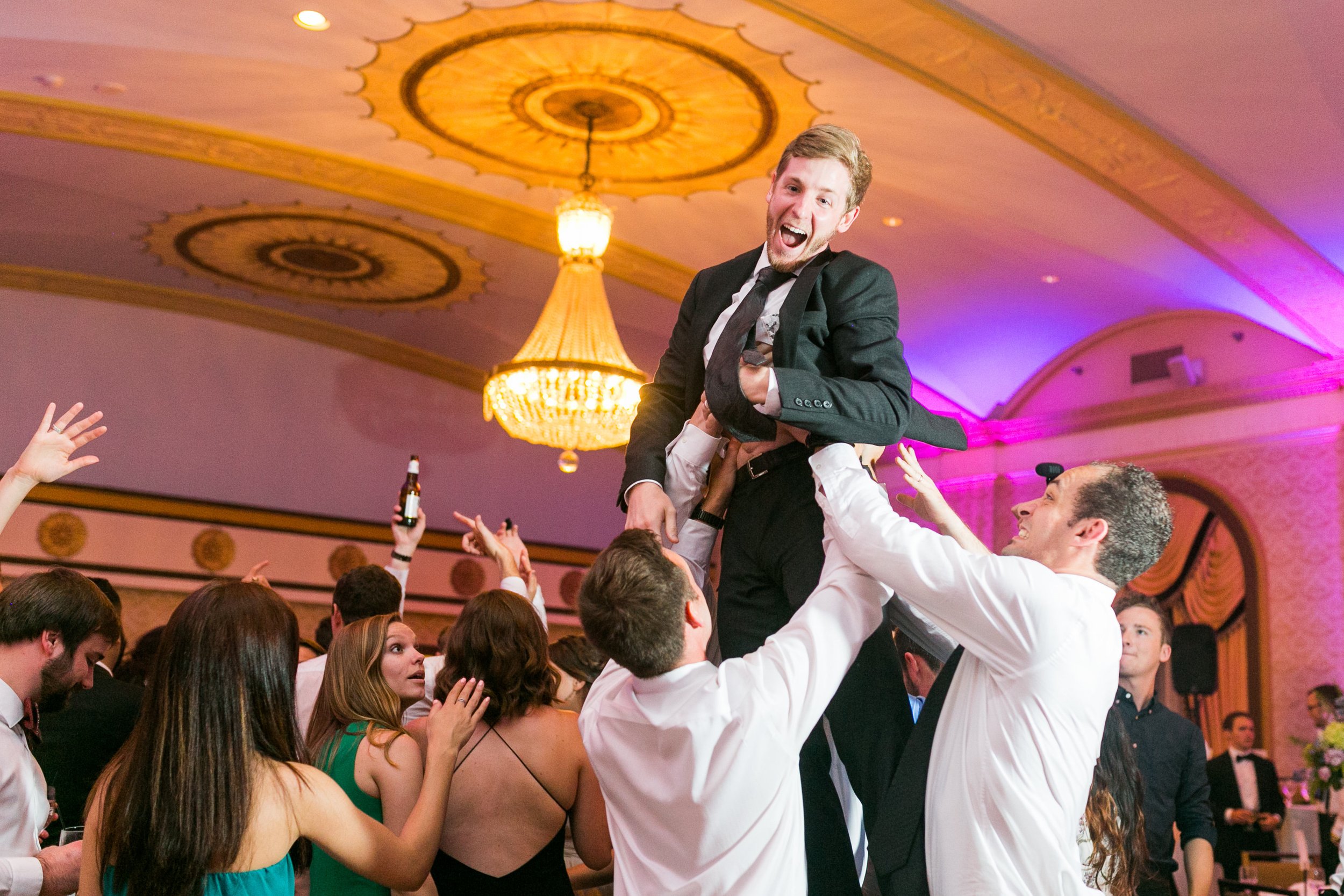 People celebrating at a wedding, lifting a man in a suit into the air inside a ballroom with a chandelier.