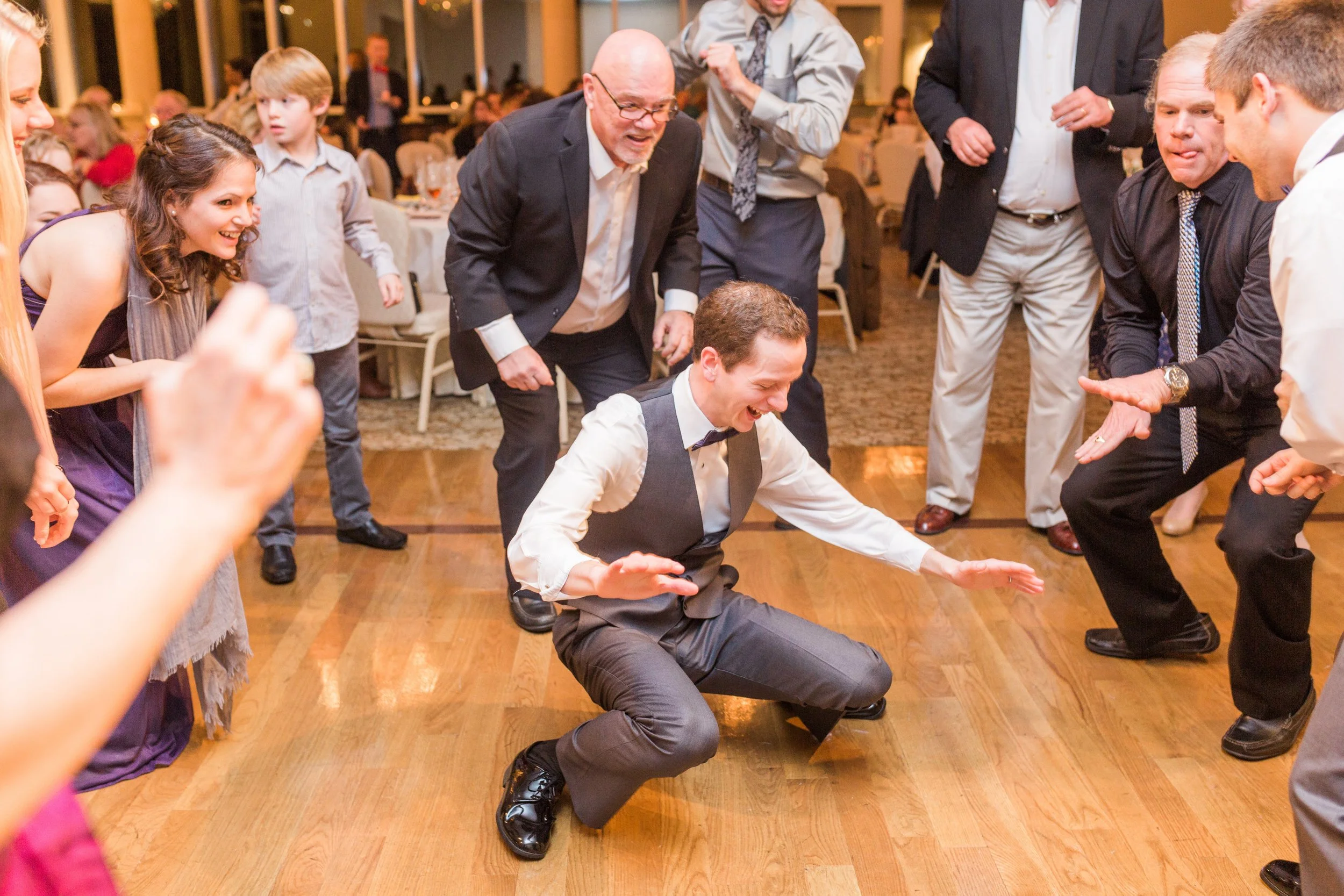 Group of people dancing and having fun at a party, with a central figure in a suit kneeling on the dance floor. The crowd is smiling and engaged, suggesting a joyous celebration, possibly a wedding or formal event.
