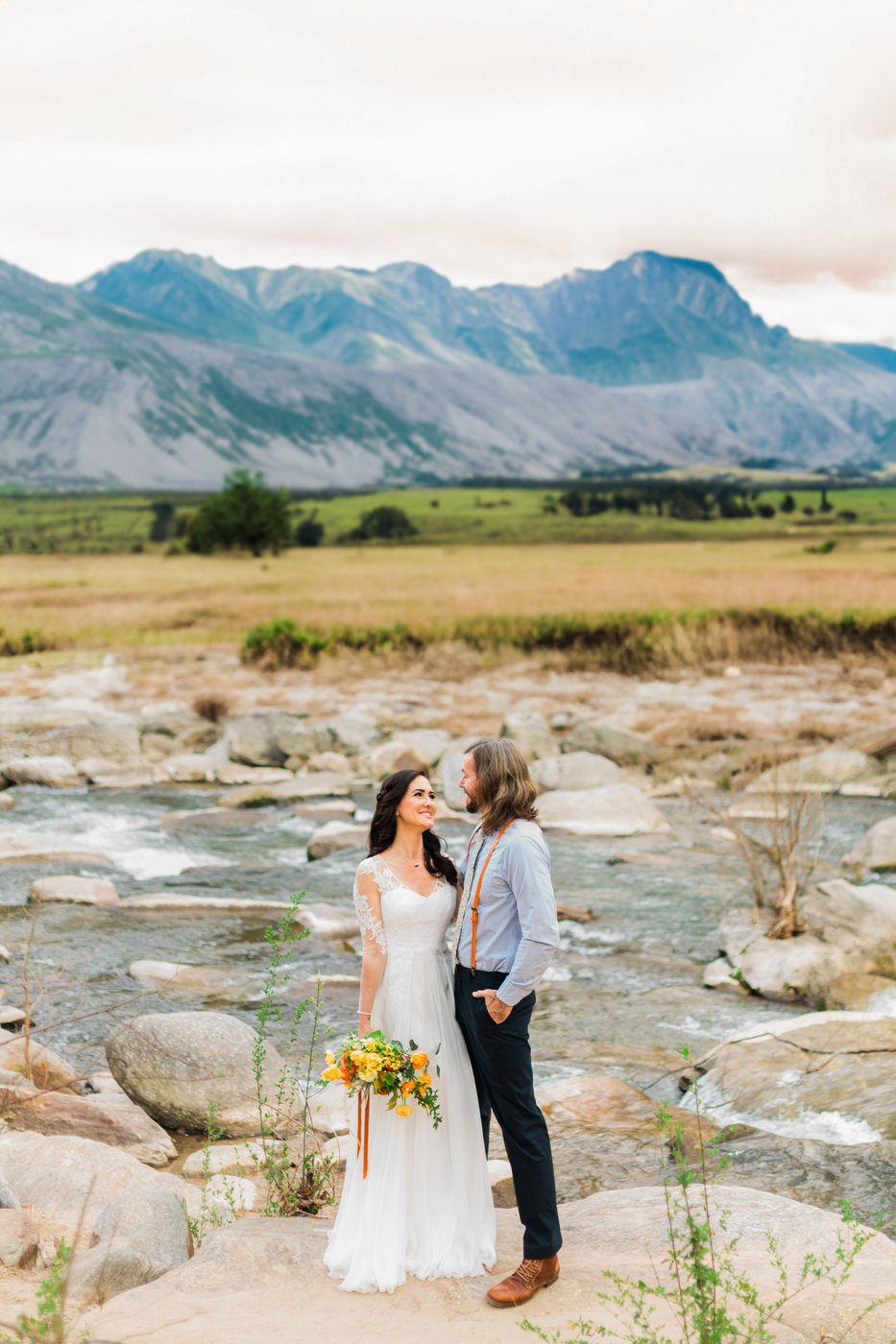 Bride and groom standing by a river with mountains in the background, wearing wedding attire, holding a bouquet of yellow flowers.