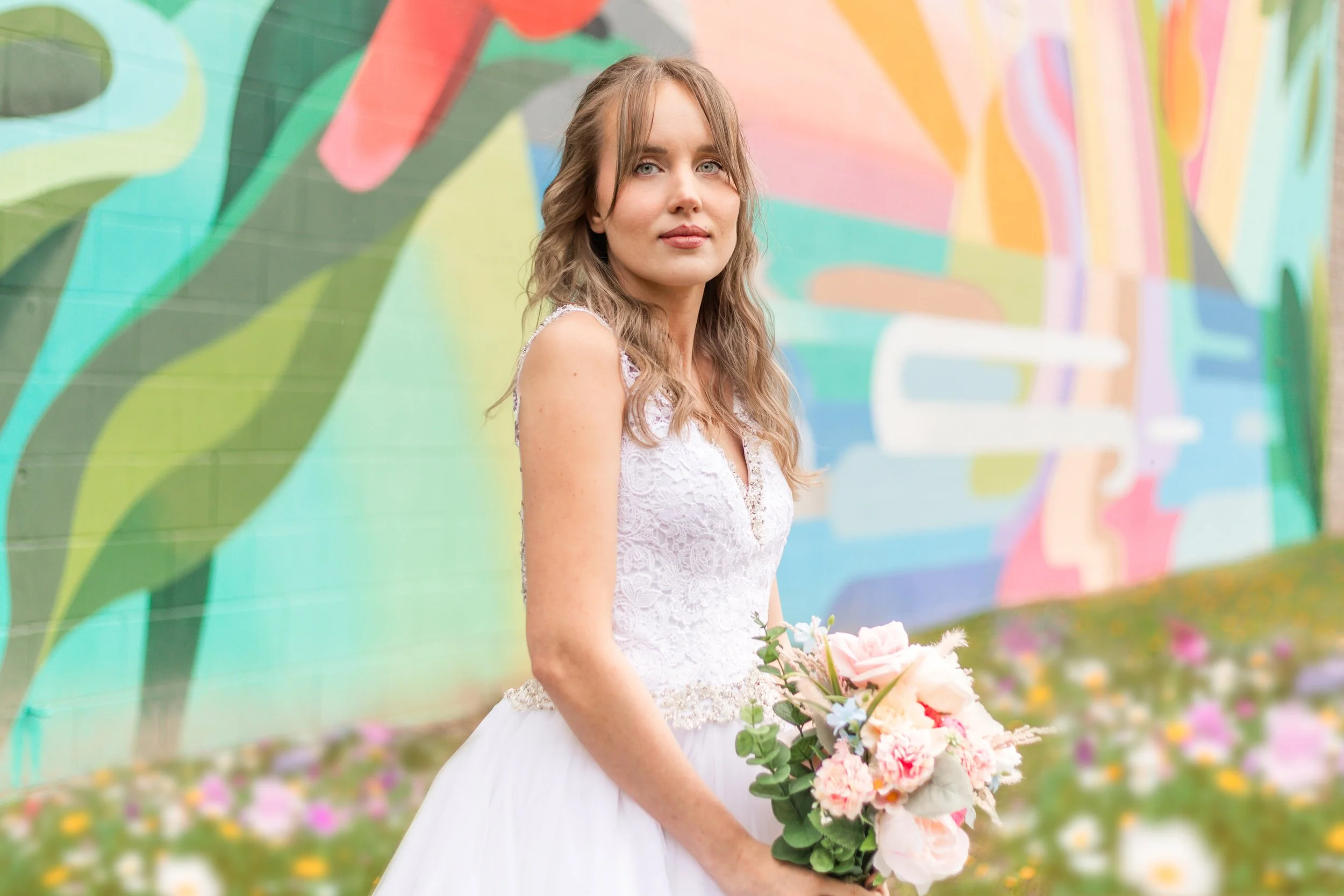 Bride in a white lace dress holding a bouquet, standing in front of a colorful mural with floral and abstract patterns.