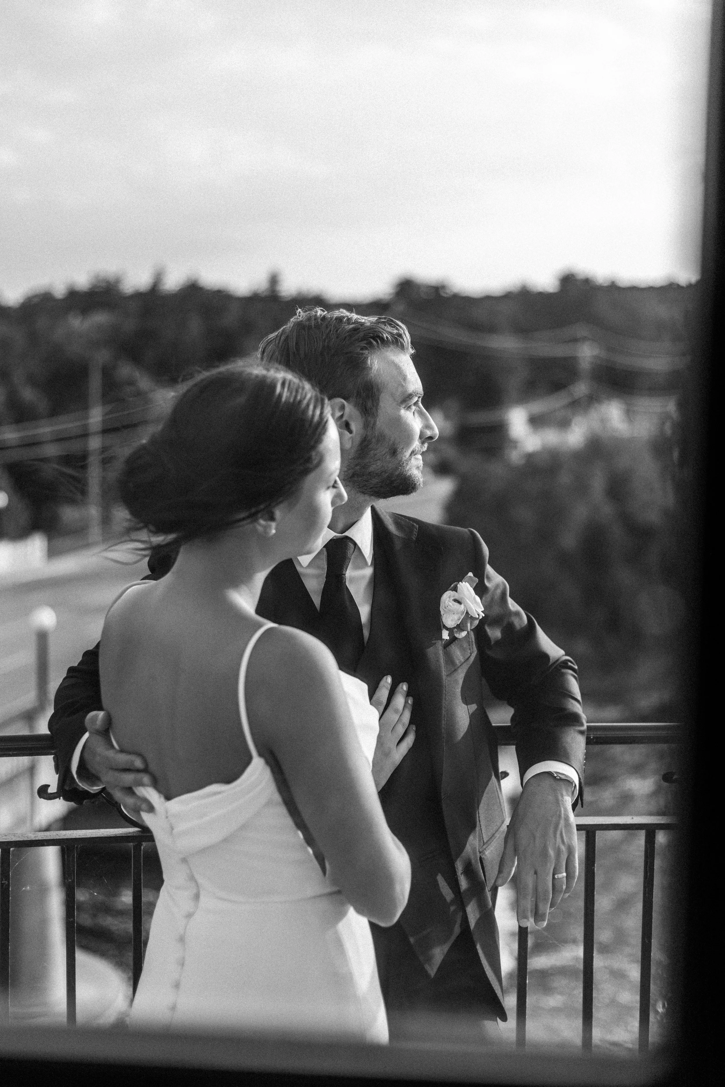 Black and white photo of a bride and groom standing on a balcony, embracing and looking into the distance, with a landscape view in the background.