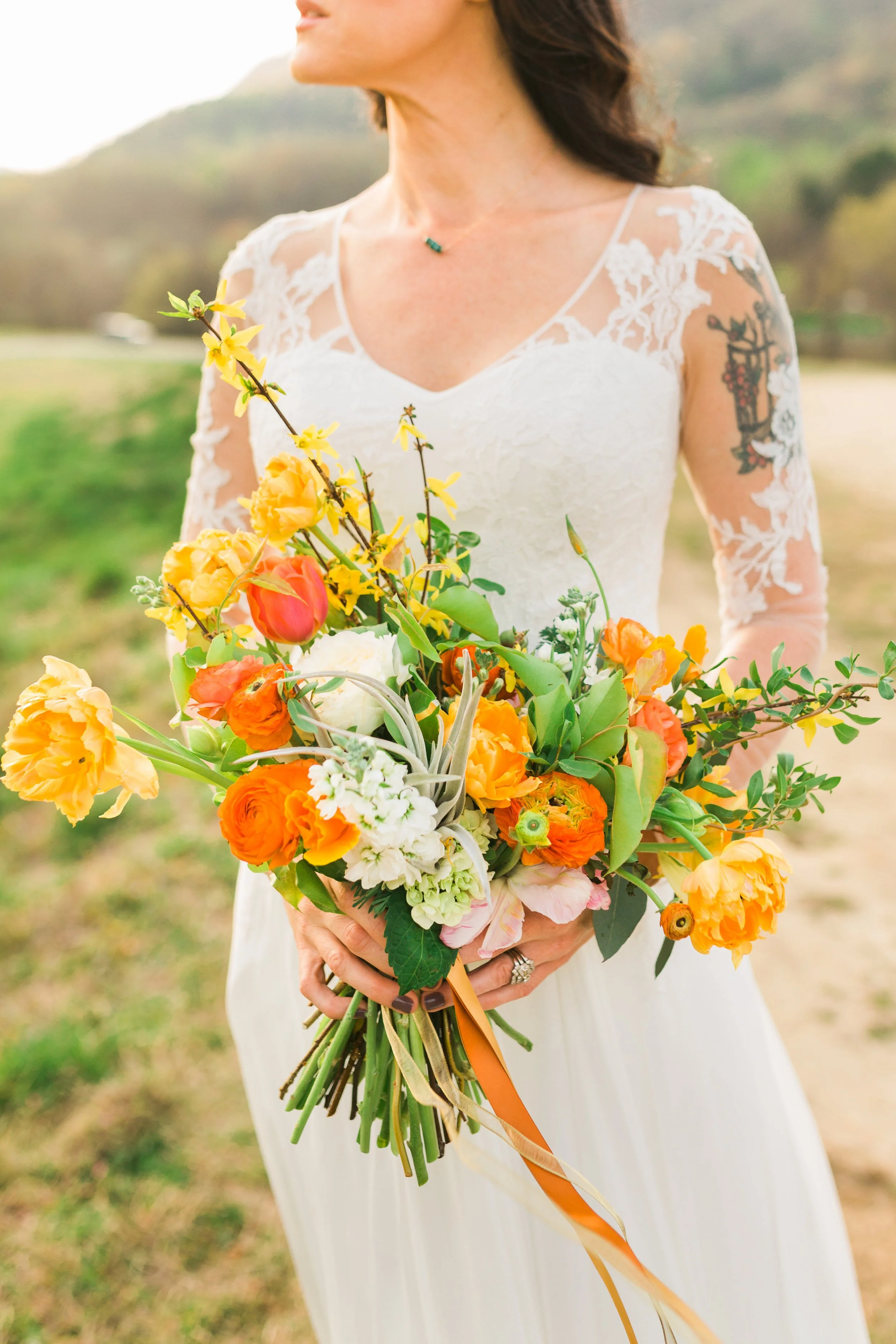 Bride holding a colorful bouquet of yellow, orange, and pink flowers with greenery, wearing a lace wedding dress in an outdoor setting.