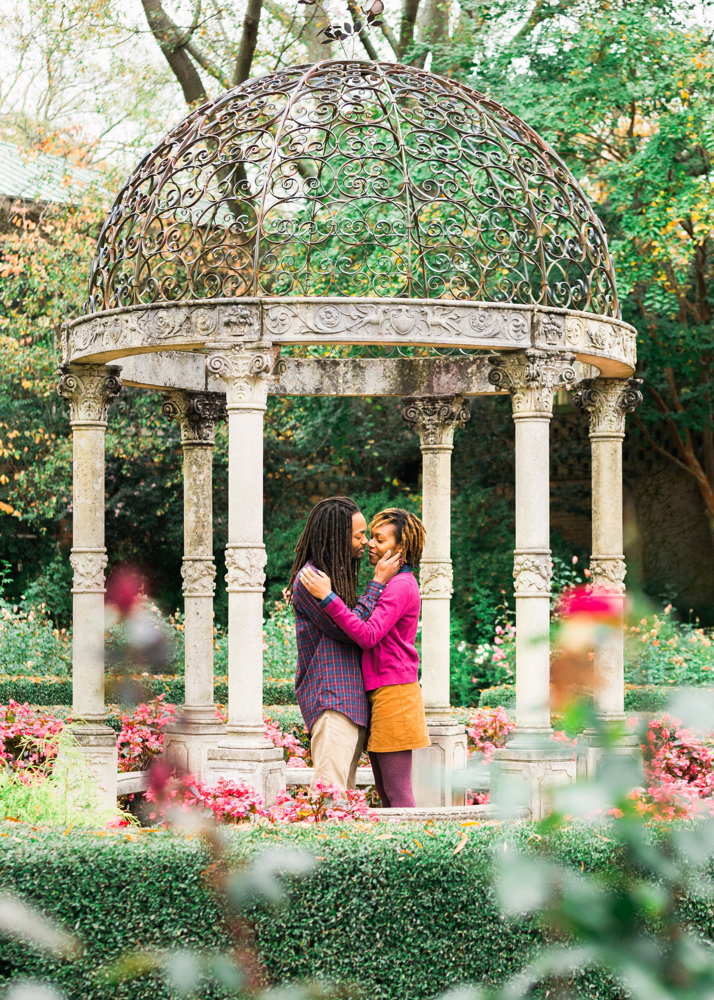 A couple embracing under a decorative stone gazebo surrounded by flowers and greenery.