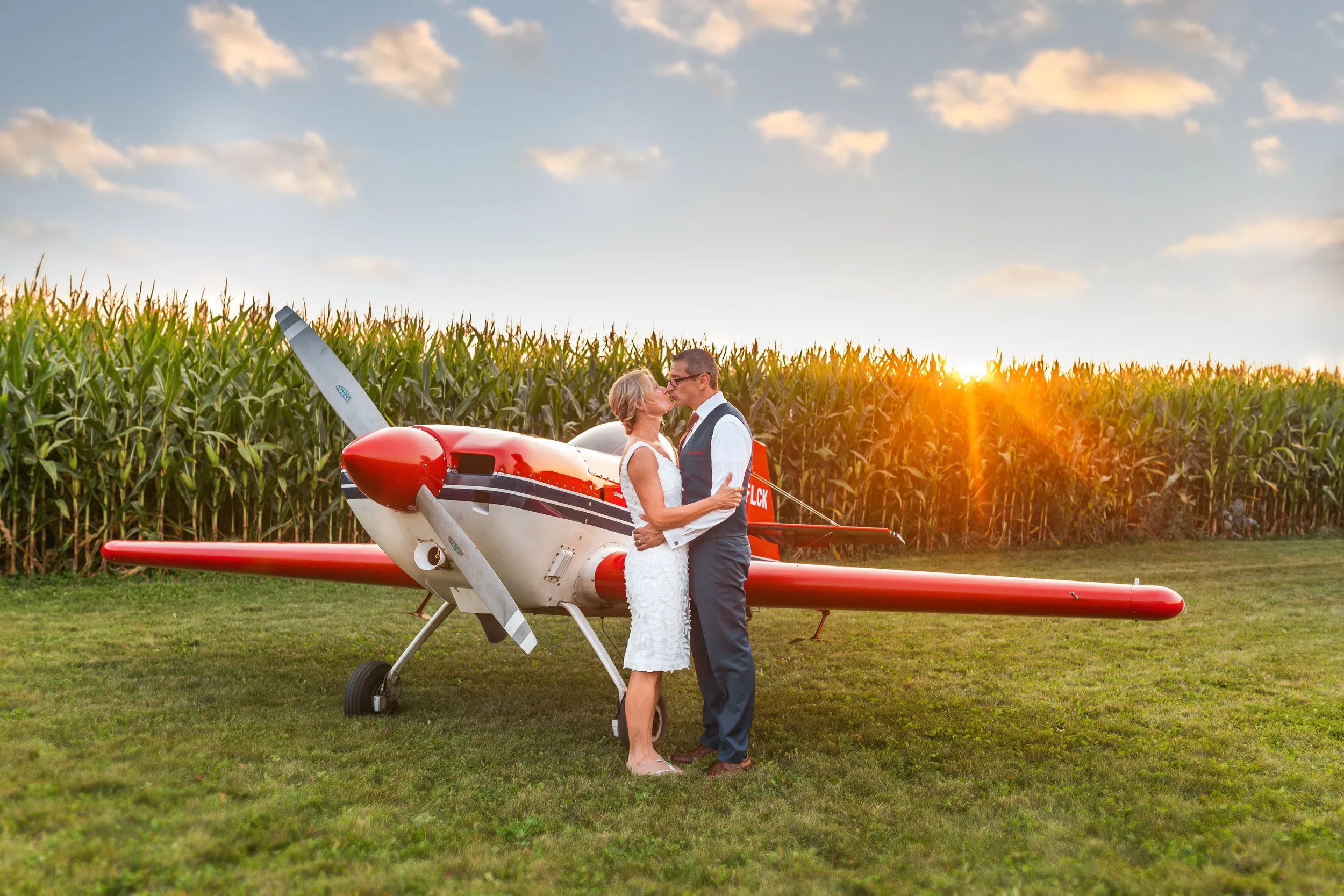 A couple kissing in front of a small red and white airplane on a grassy field with a cornfield and a sunset in the background.