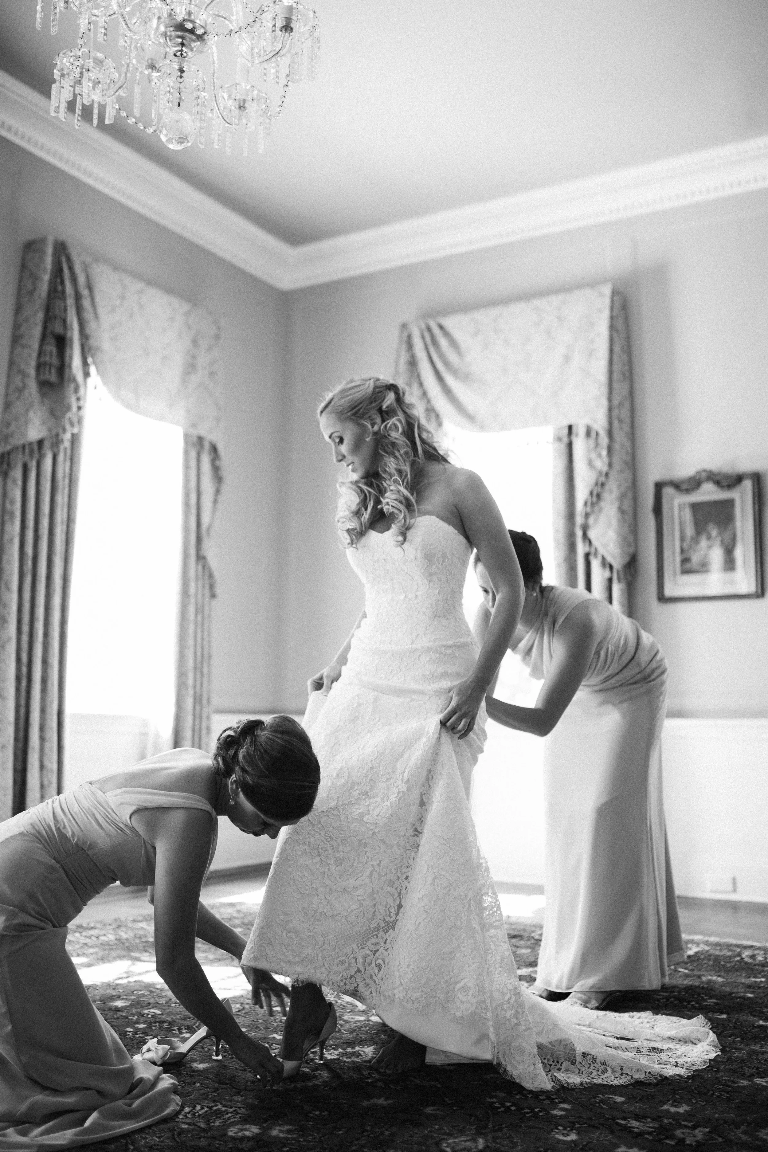 Bride in a wedding dress being helped by two women; one kneeling to adjust shoes, the other adjusting the dress, in a room with elegant decor.