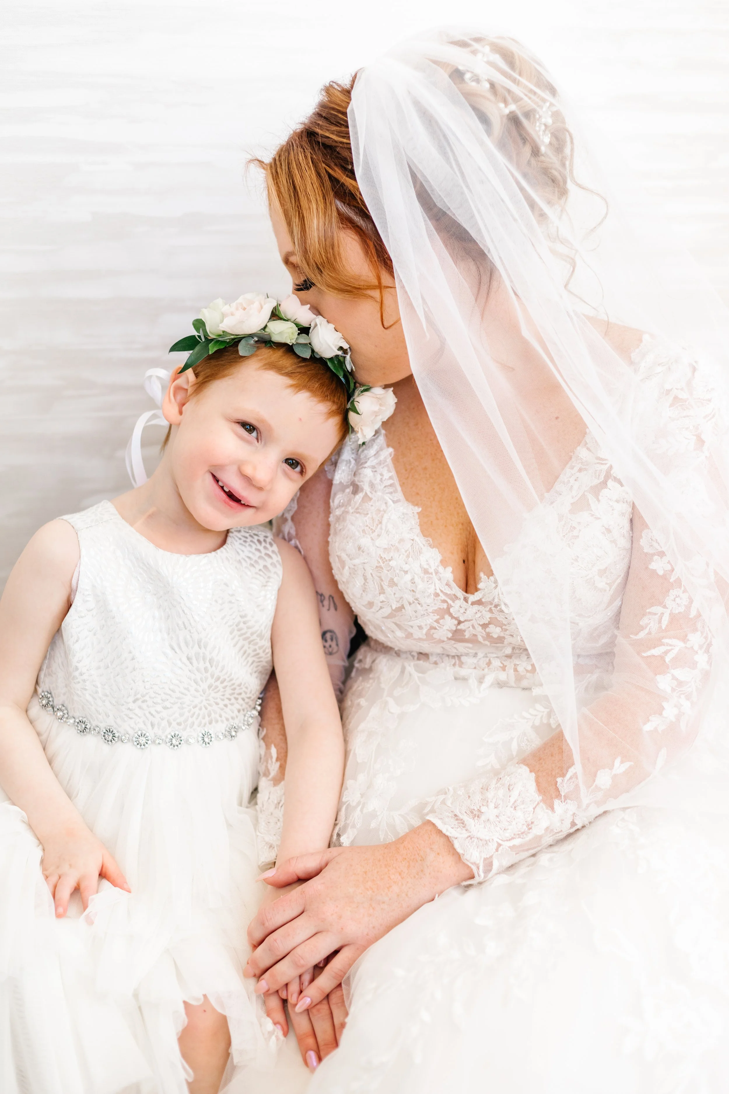 Bride and young girl in white dresses, the girl wearing a floral crown and the bride in a veil, sitting closely together.