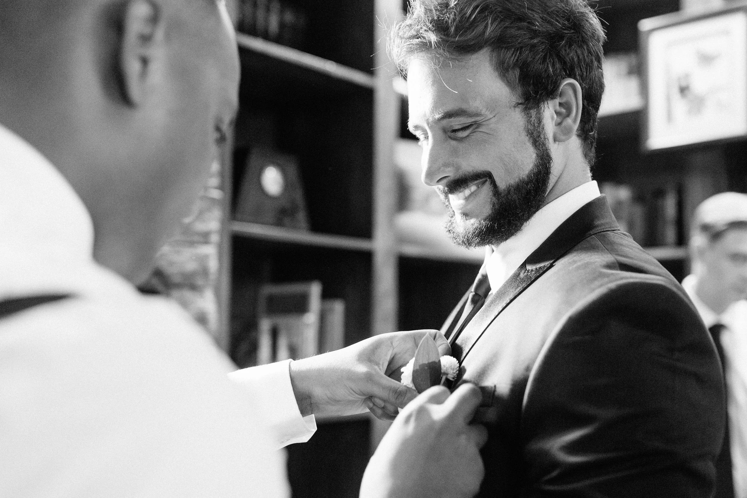 Two men in formal attire, one smiling while the other adjusts his boutonnière, in a room with bookshelves, in a black and white image.