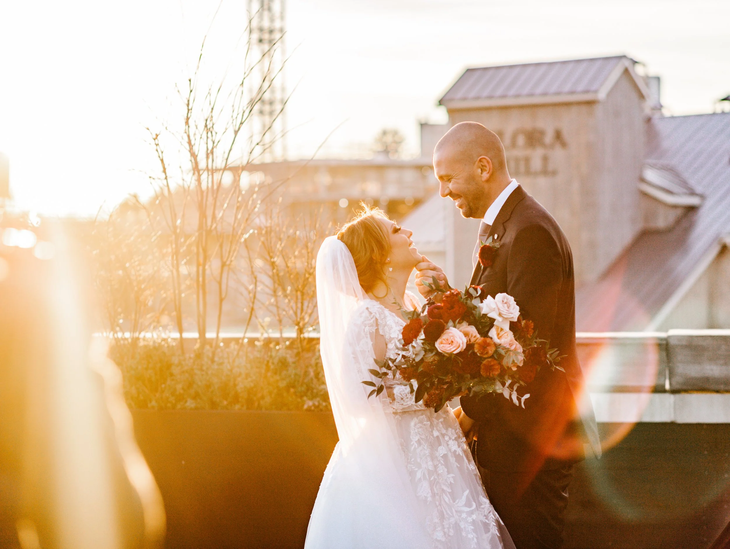Bride and groom smiling at each other during sunset, holding a bouquet with roses, on a rooftop.