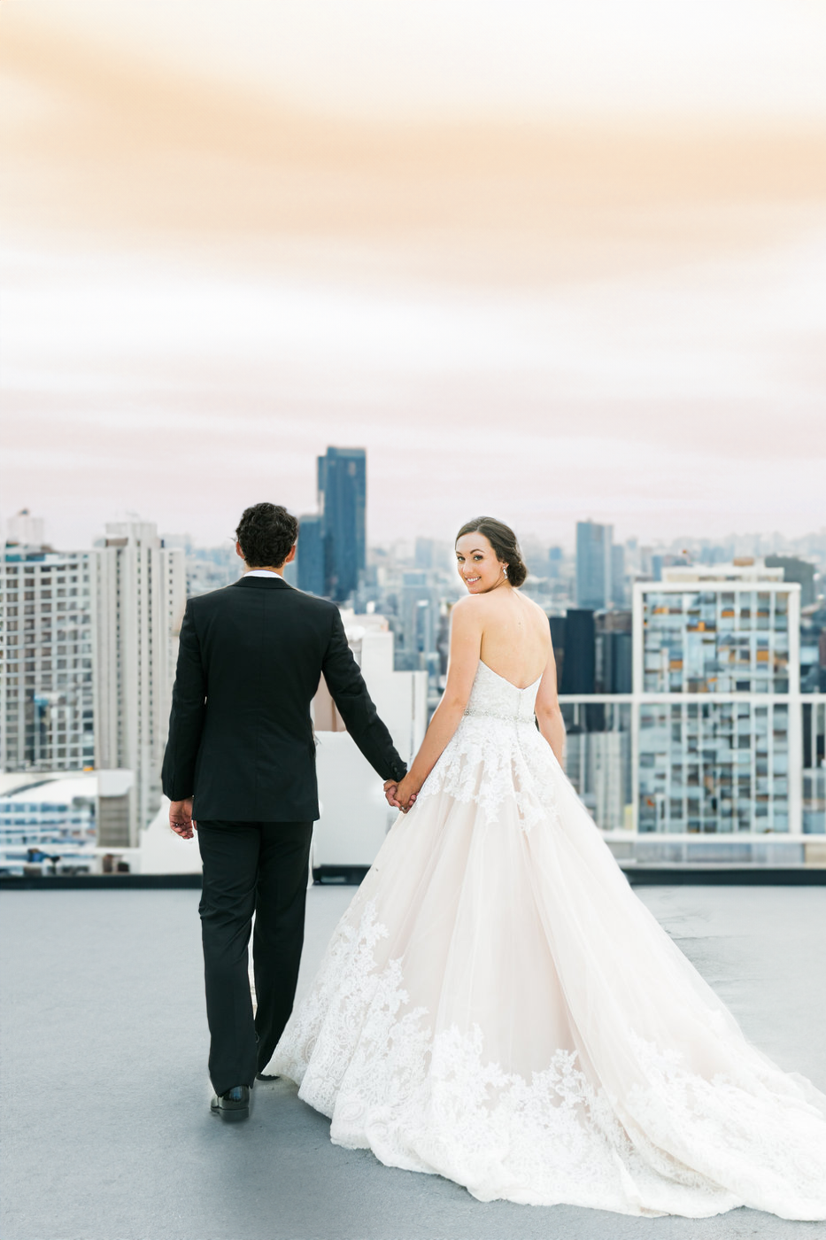 Bride and groom holding hands on a rooftop with a city skyline in the background.