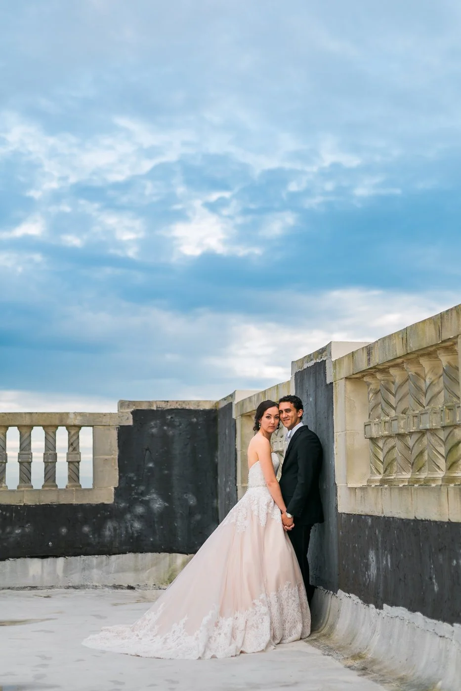 Bride and groom standing on a rooftop with stone railings, holding hands and looking at the camera, under a cloudy blue sky.