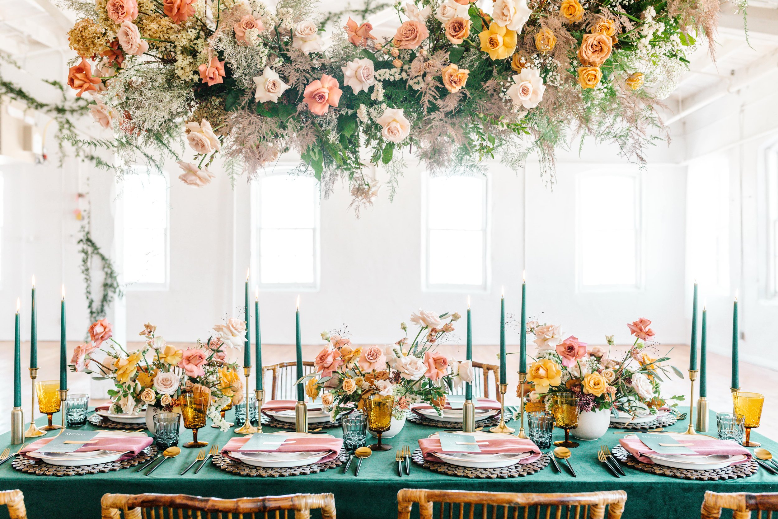 A beautifully set dining table decorated for a formal event. The table features pink napkins, gold cutlery, and elegant floral centerpieces with pink, peach, and yellow roses. Blue-green candles in gold holders add a touch of elegance. Above the tabl