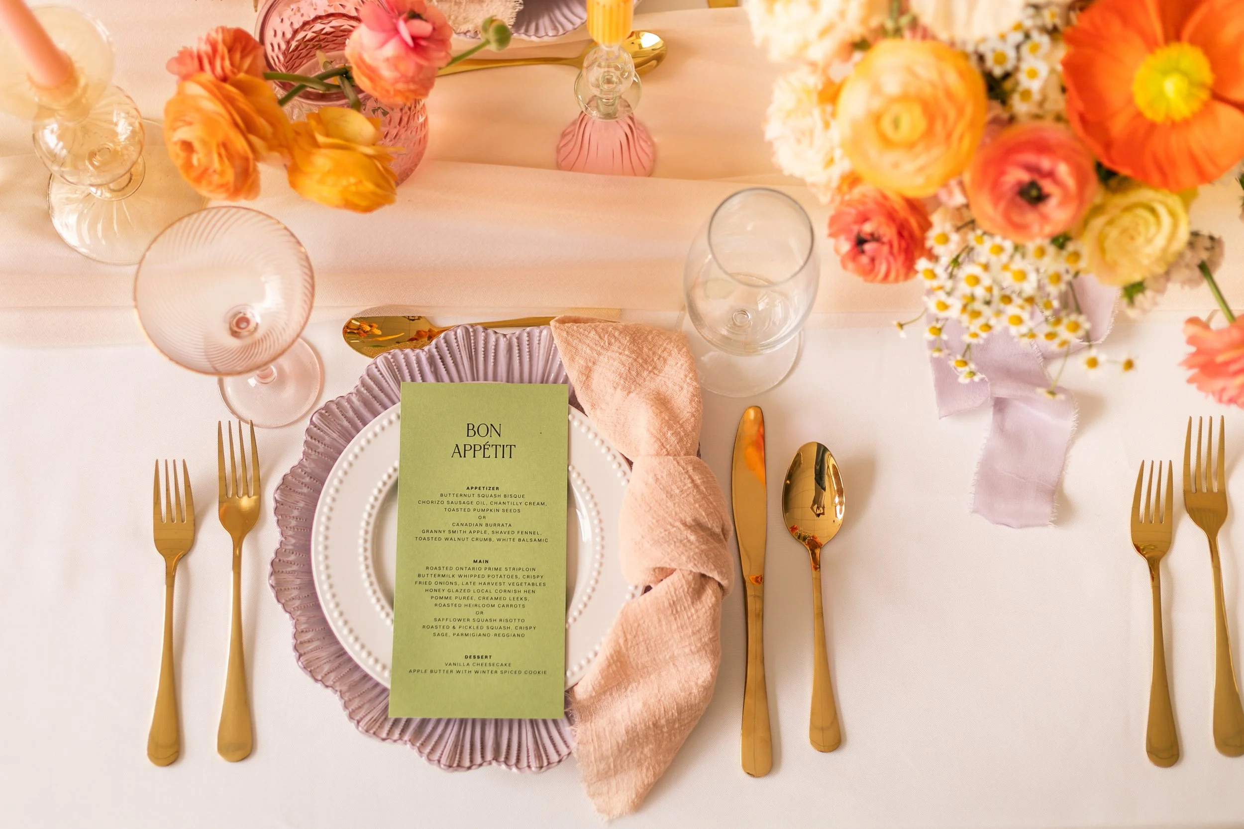 Elegant table setting with a green menu card, pink napkin, gold utensils, and floral arrangement.