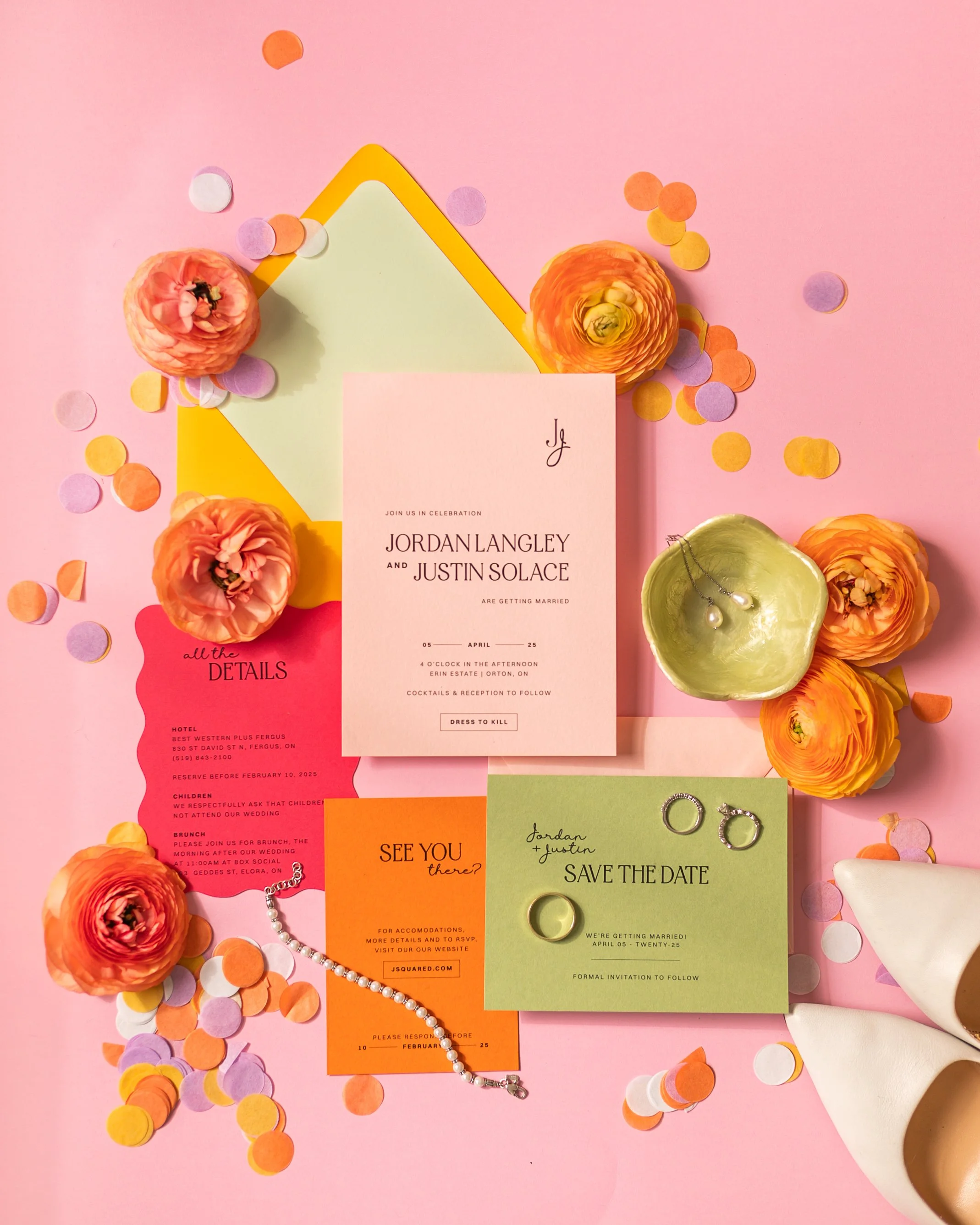 Wedding invitation suite on pink background with flowers, envelopes, confetti, and wedding rings.