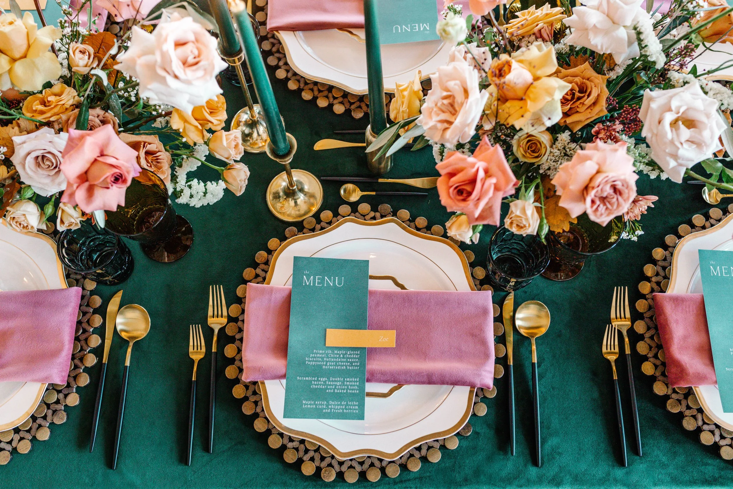 Elegant dining table setting with plates, pink napkins, menus, gold cutlery, floral centerpiece, and green candles on a dark green tablecloth.