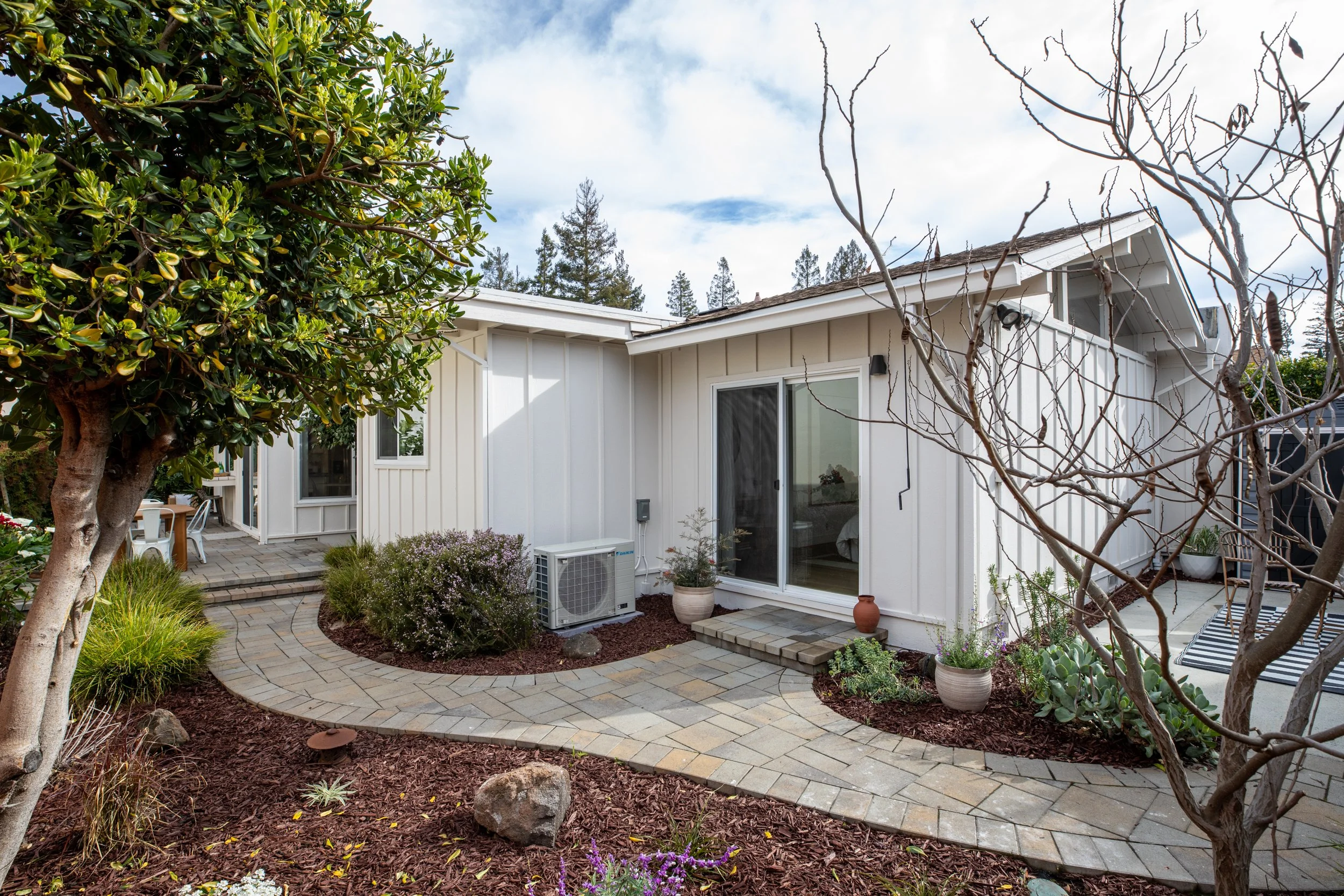 View of a modern house with white exterior walls, a sliding glass door, potted plants, a curved stone pathway, and surrounding trees and shrubs in a landscaped yard.