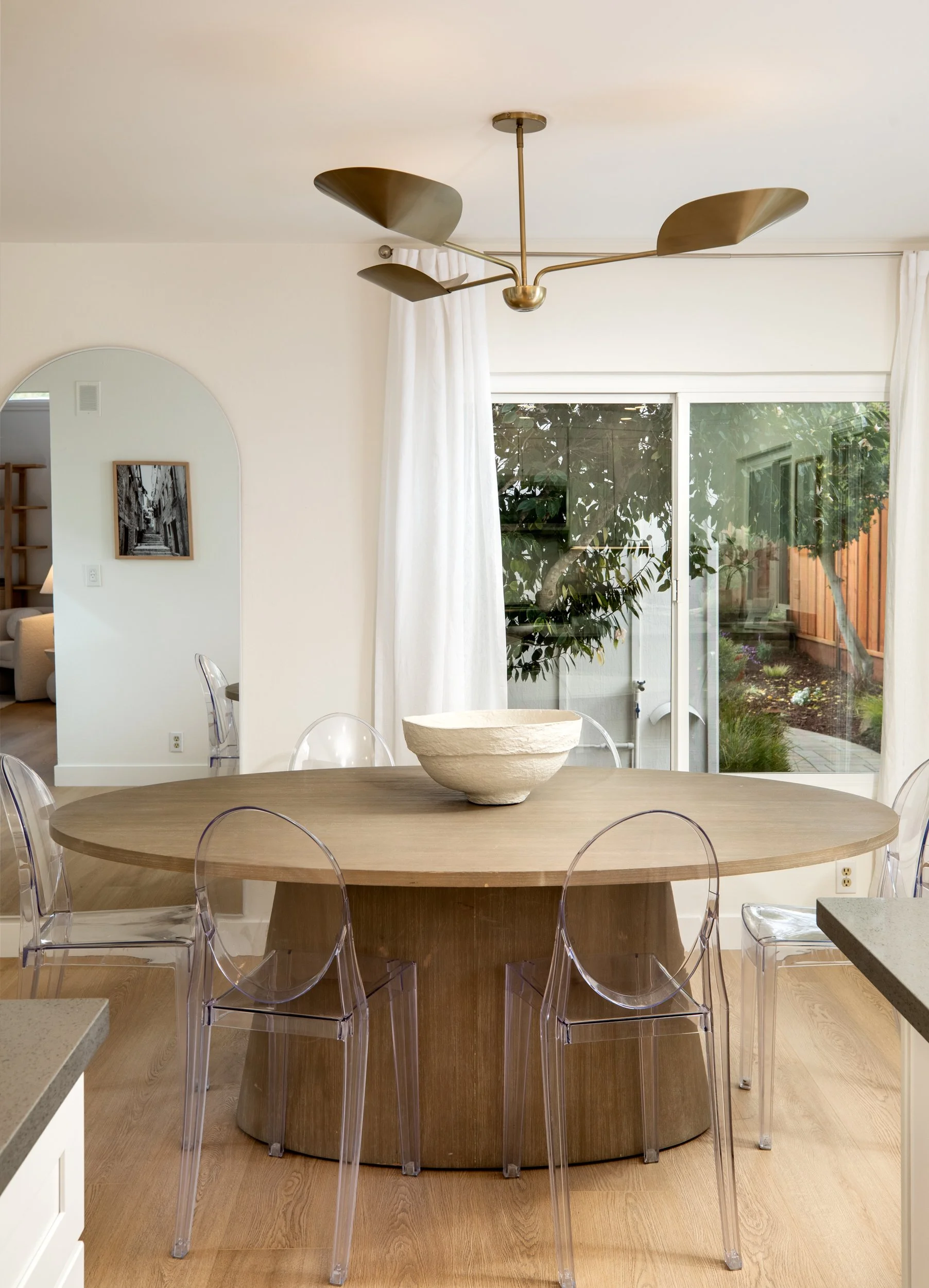 A dining room with a round wooden table, transparent ghost chairs, a large ceramic or stone bowl in the center, and a modern gold-colored ceiling lamp. There is a large sliding glass door with white curtains, overlooking a backyard with a tree and a 