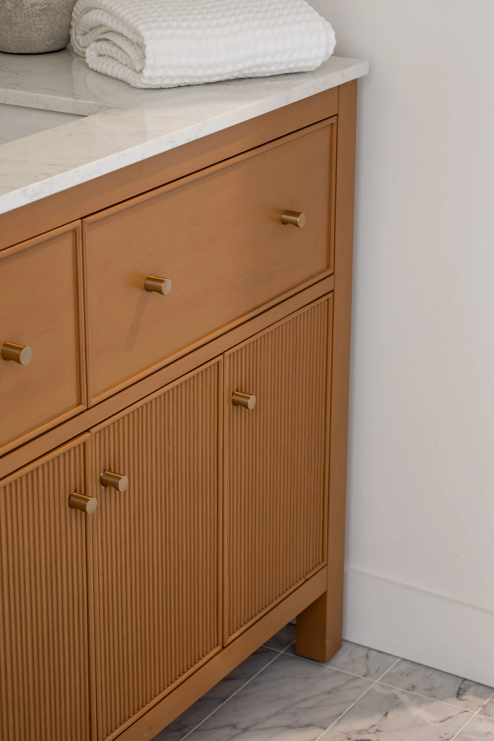 Close-up of a wooden bathroom cabinet with grooved doors, gold knobs, and a white marble top with folded white towels on it.