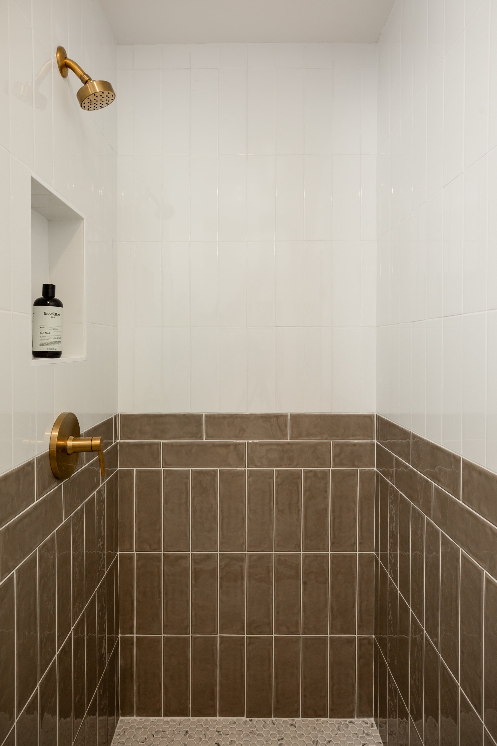 Empty walk-in shower with white tile upper walls, brown tile lower walls, a gold showerhead, a gold faucet, and a built-in shelf with a black bottle inside.