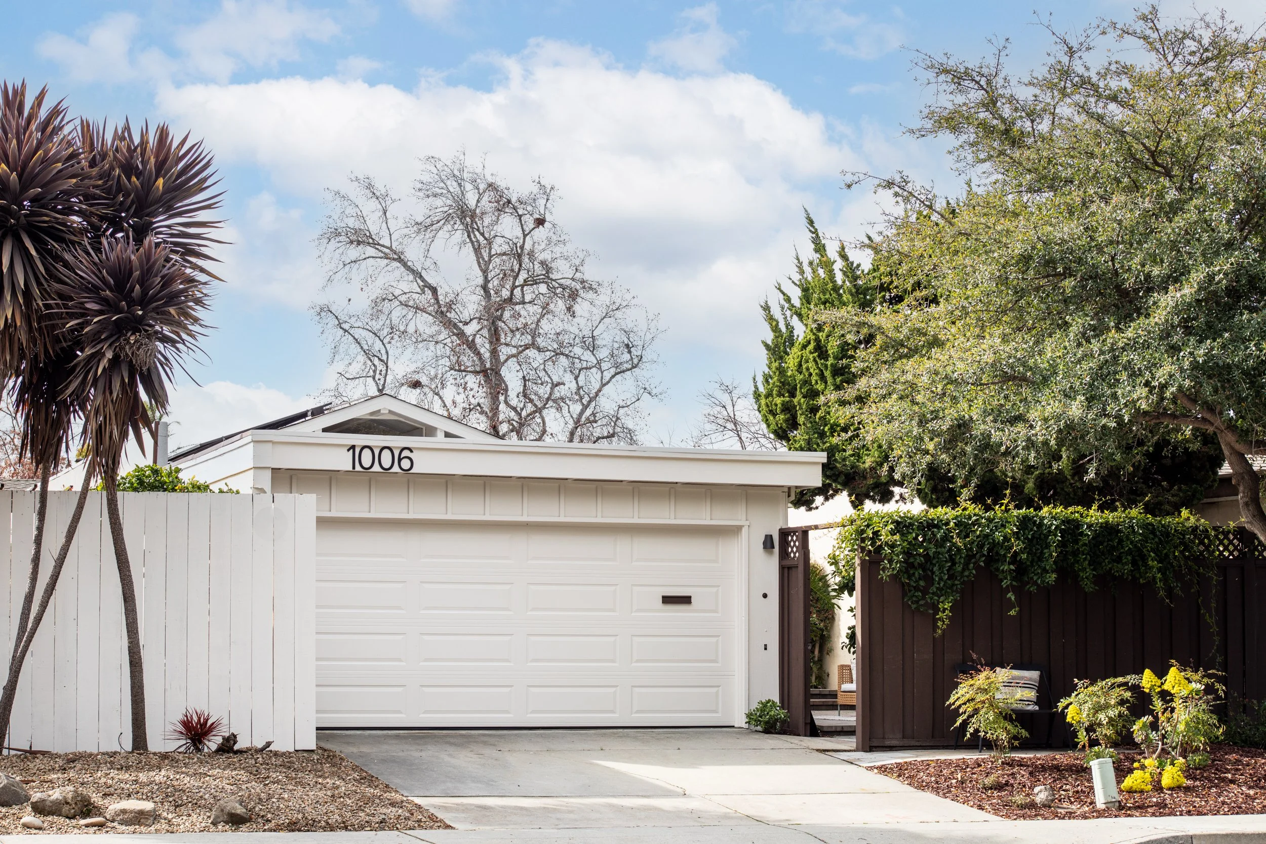 Front view of a modern residential house with white garage door and house number 1006, surrounded by trees and bushes, with a rock garden in the front yard and a sidewalk.