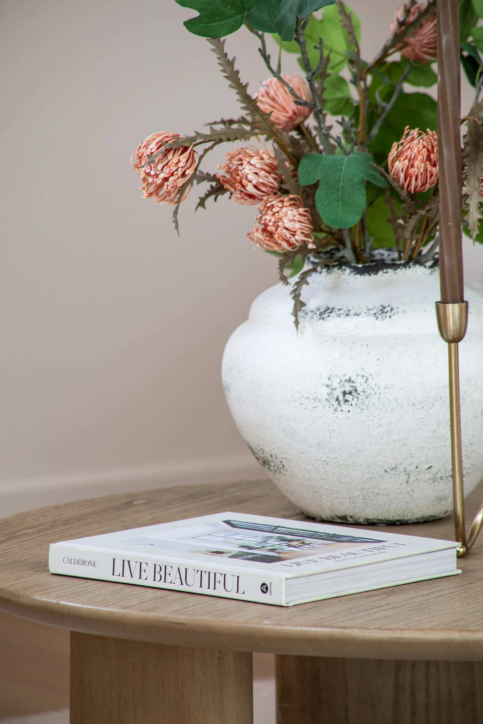 A white textured ceramic vase filled with pink and orange flowers and green leaves, placed on a wooden table next to a closed book titled "Live Beautiful".