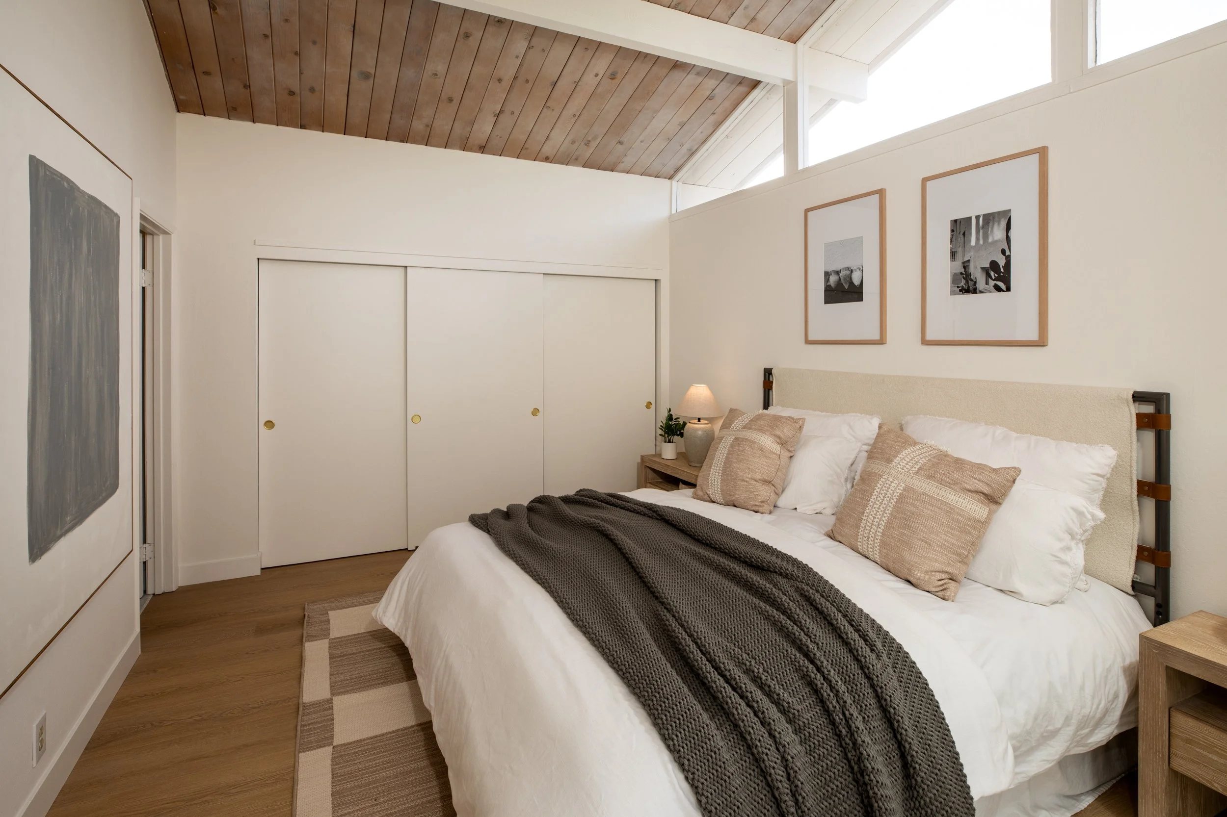 A cozy bedroom with a large bed, beige and white pillows, a dark gray knitted throw blanket, a wooden bedside table with a lamp and plant, three black and white framed photos on the wall, wooden closet doors, and a wood-paneled ceiling with high wind