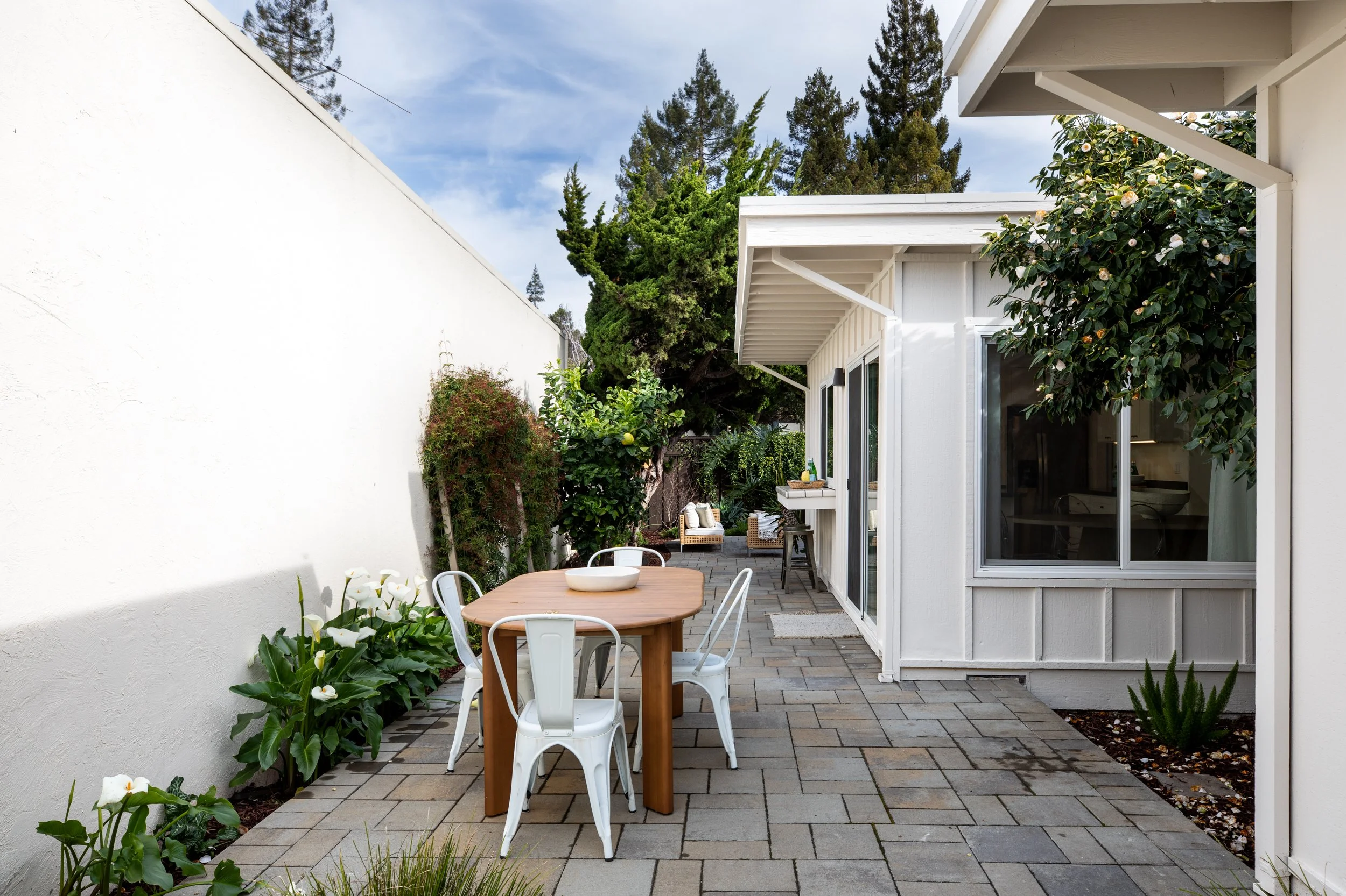 Outdoor patio with a wooden table and white metal chairs, surrounded by plants and trees, next to a white house with sliding glass doors.