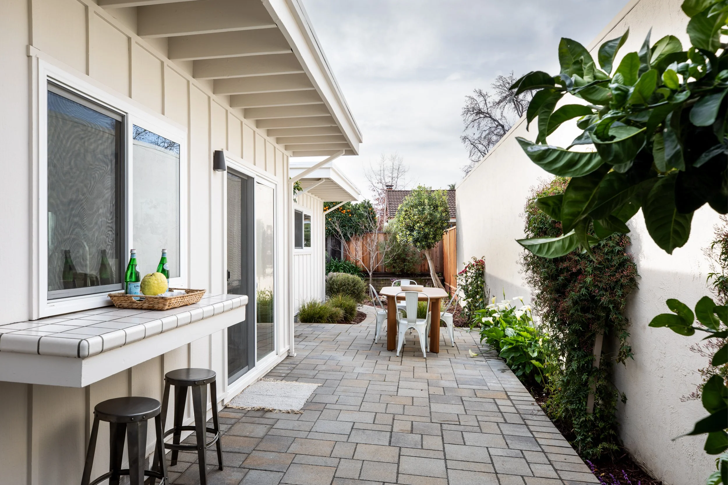 A backyard patio with potted plants, a small round table with four white chairs, and a white wall on the right. The house has white exterior walls, sliding glass door, and window, with a small tiled bar area with two stools on the left. The patio is 