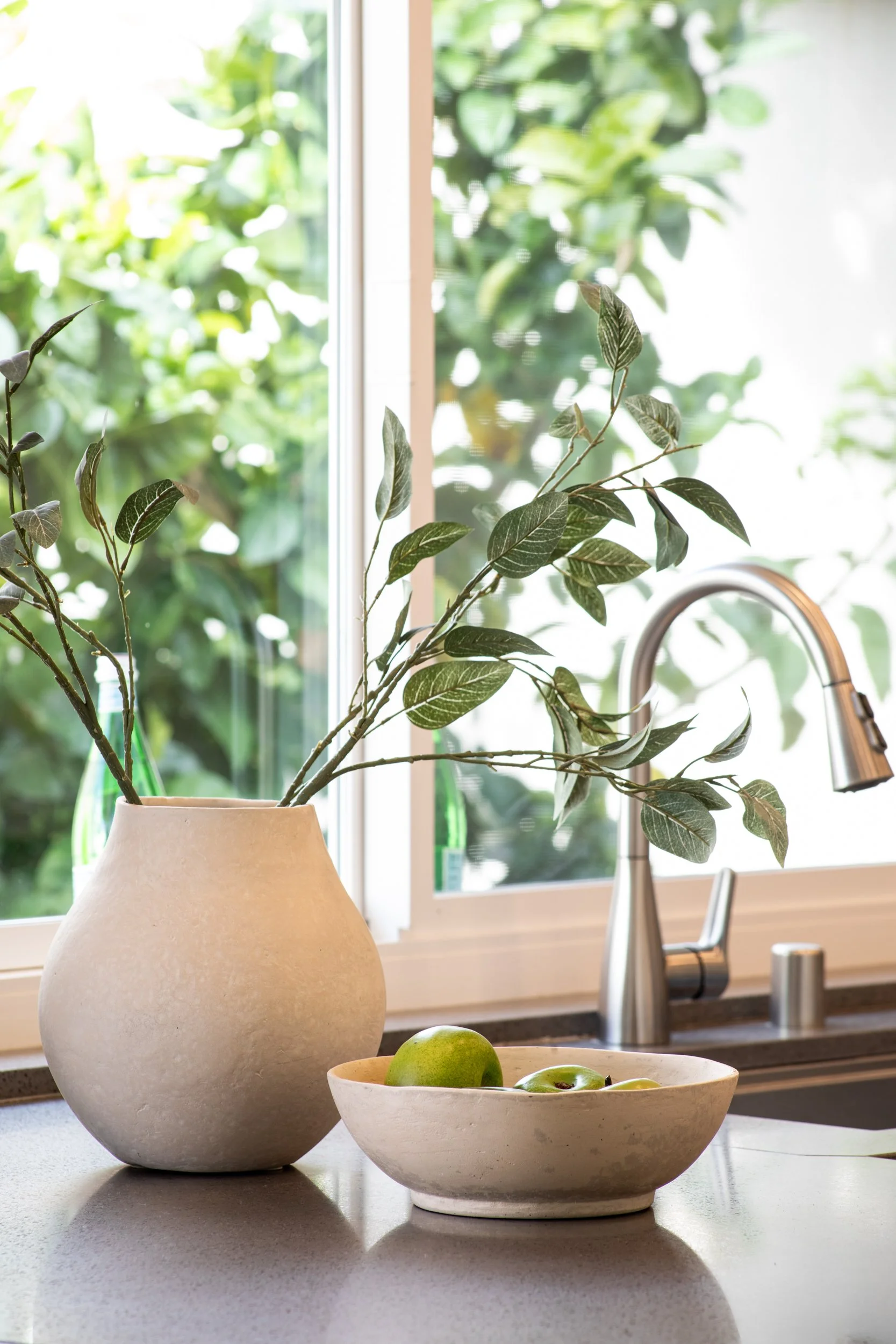 A kitchen counter with a beige vase holding leafy branches, a white bowl with green apples, and a stainless steel sink and faucet near a window, with greenery outside.