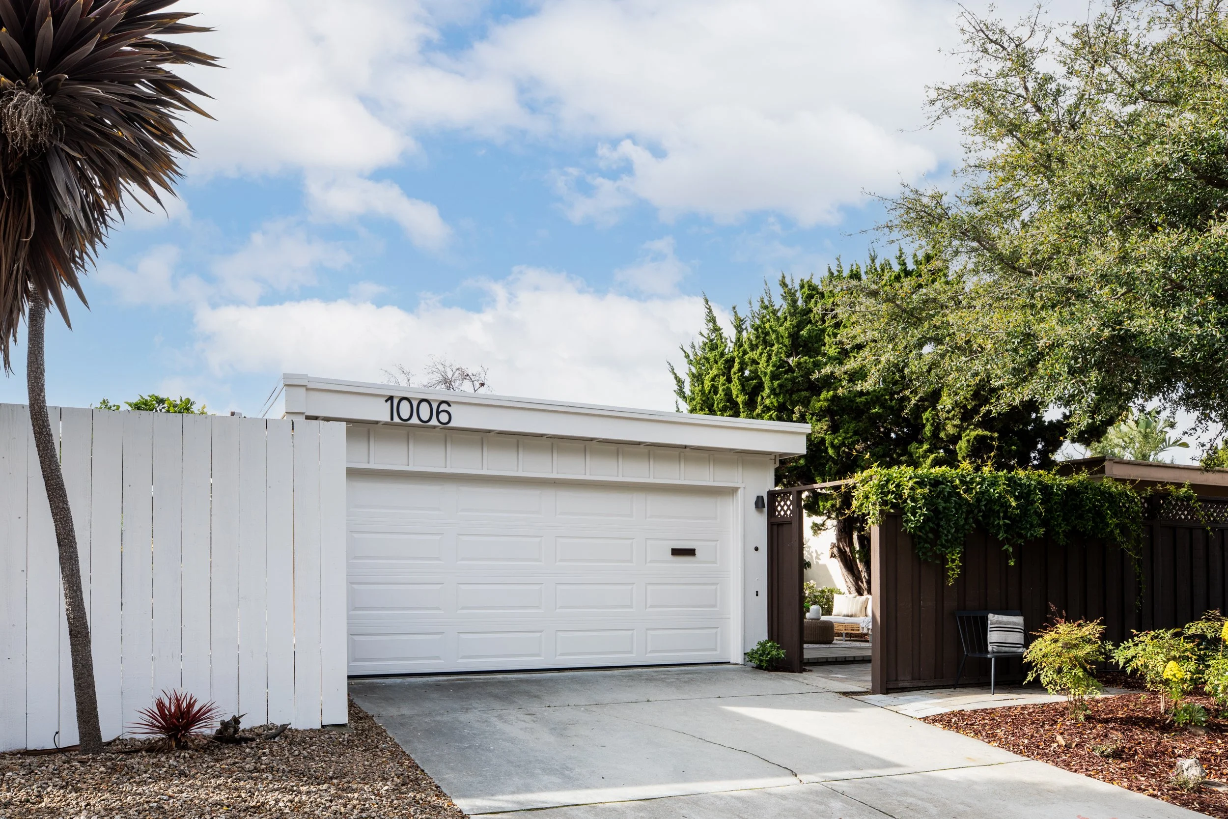 Front view of a modern house with a white garage door, a white fence on the left, and a small patio area on the right, with trees and shrubs around.