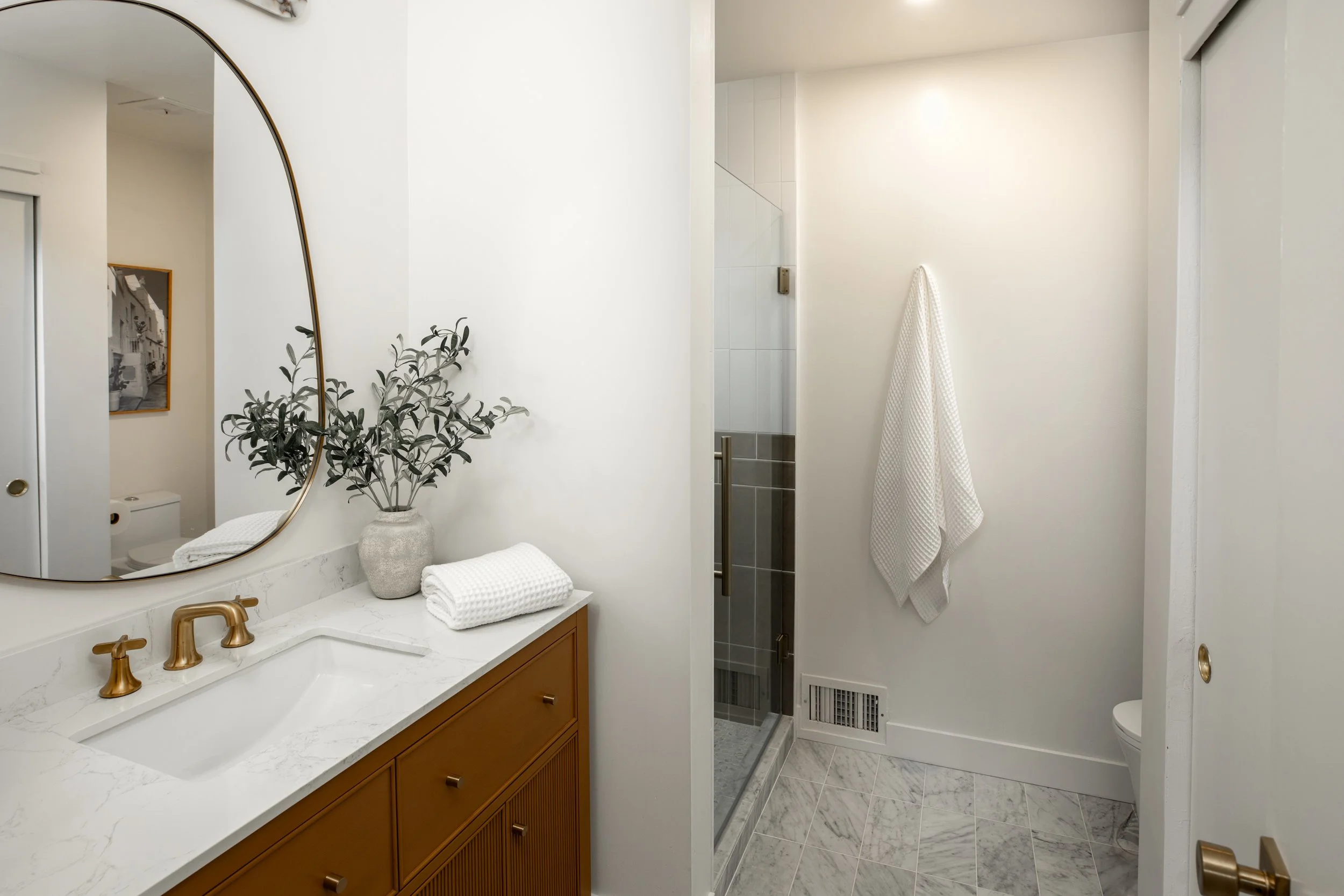 Bathroom with a white marble countertop, round mirror, green plant in a vase, folded white towels, a shower with a glass door, white waffle-weave towel hanging on the wall, toilet partially visible, and marble tile flooring.
