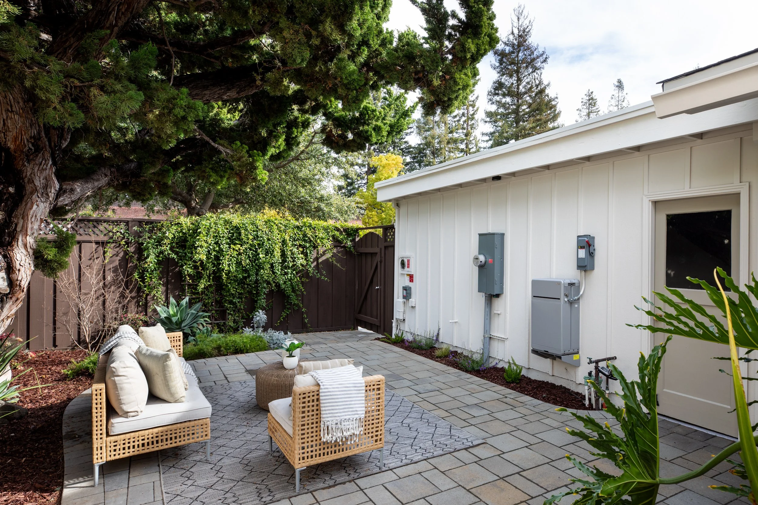 A cozy outdoor patio area with beige wicker furniture, pillows, a small table with a plant, all on a patterned rug, surrounded by plants, trees, and a white house wall with utility meters.