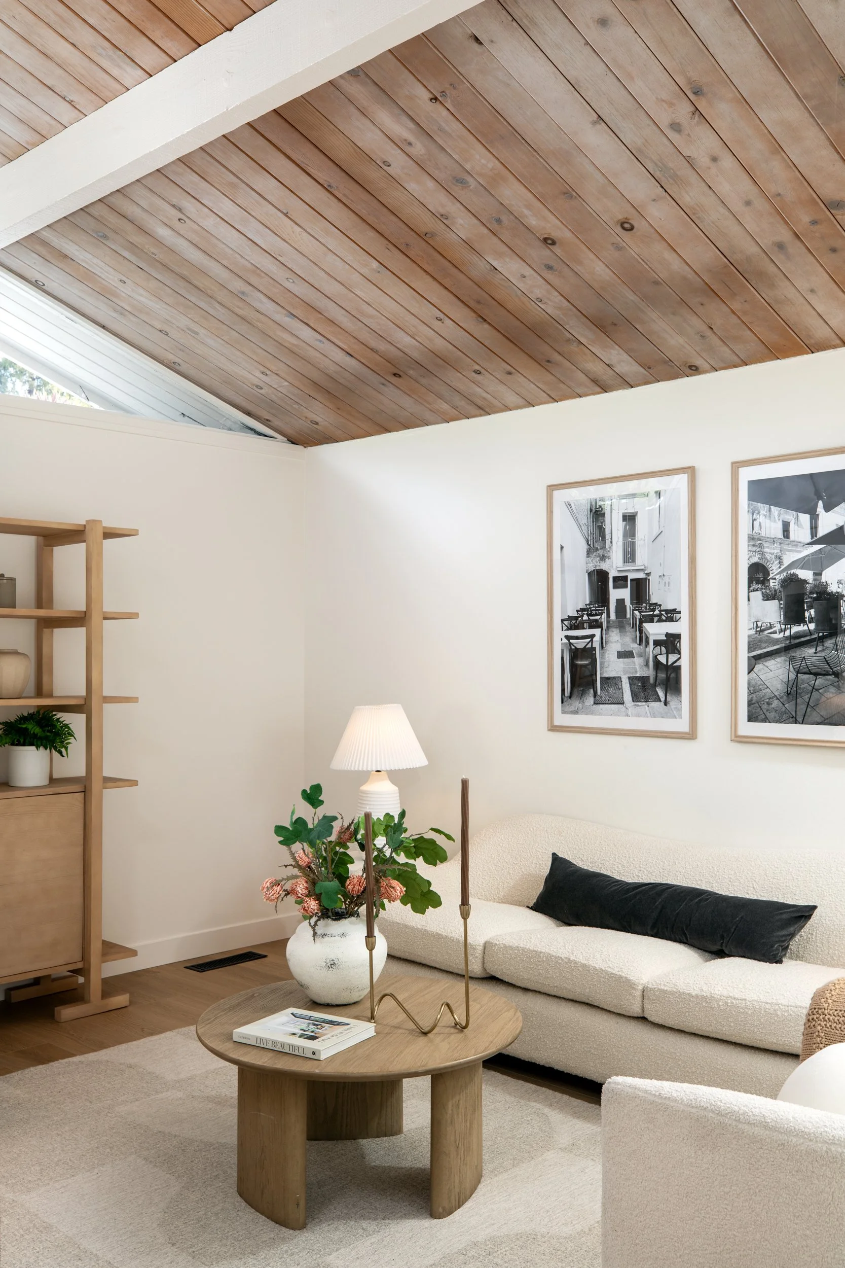 Living room with wooden ceiling, white walls, cream sofa with black pillow, wooden side table with vase of pink flowers, white table lamp, and framed black-and-white photographs on the wall.