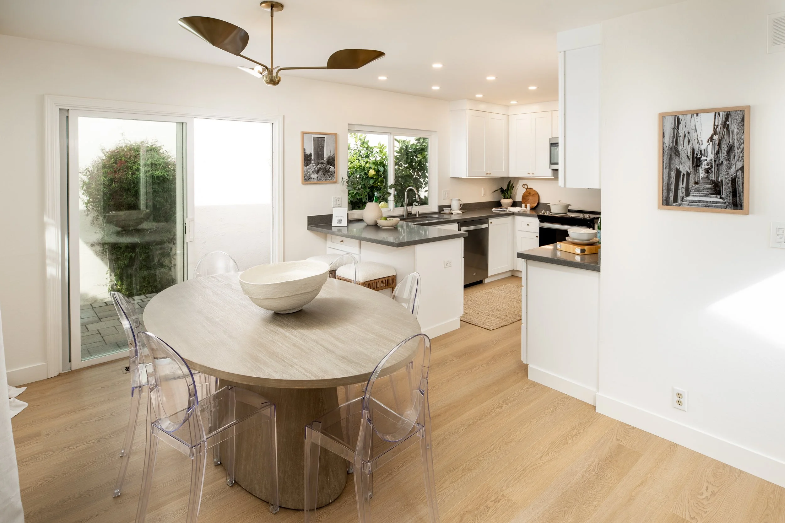 Bright modern kitchen and dining area with a round wooden table, transparent chairs, white cabinetry, and sliding glass door leading outside.