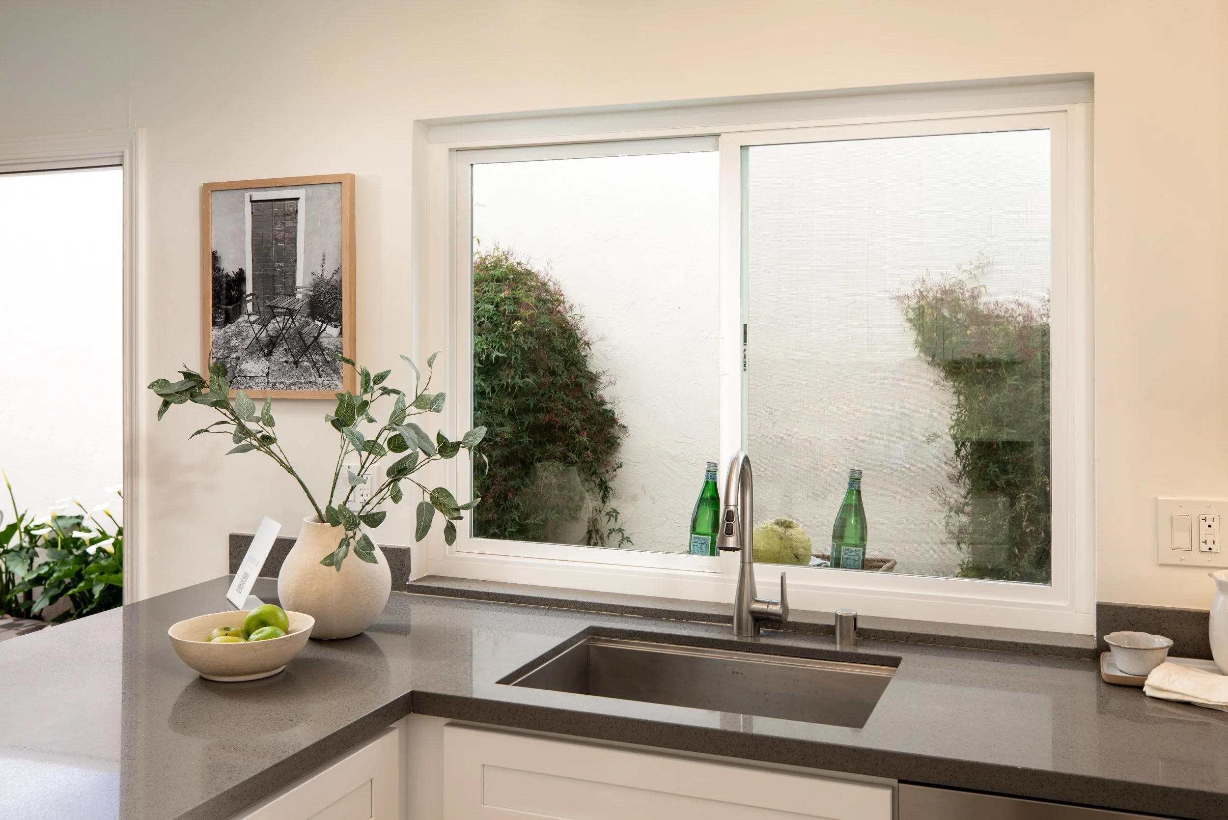 Kitchen sink with a window overlooking a small outdoor patio with bushes, two glass bottles, and a green fruit.