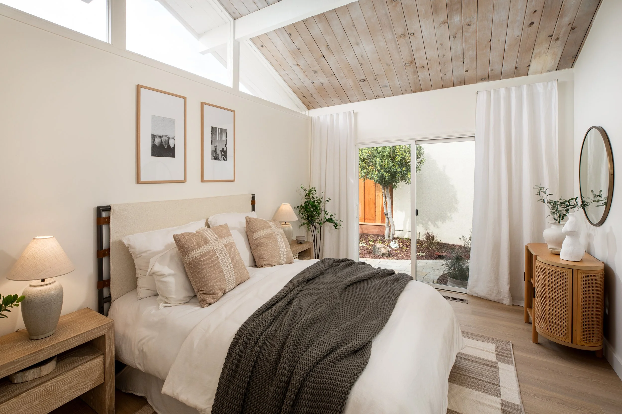 A cozy bedroom with a bed covered in white linens and beige pillows. There are wooden nightstands with lamps on each side of the bed, and a wooden dresser with vases and plants on it. The room has a vaulted ceiling with wood paneling, large windows, 