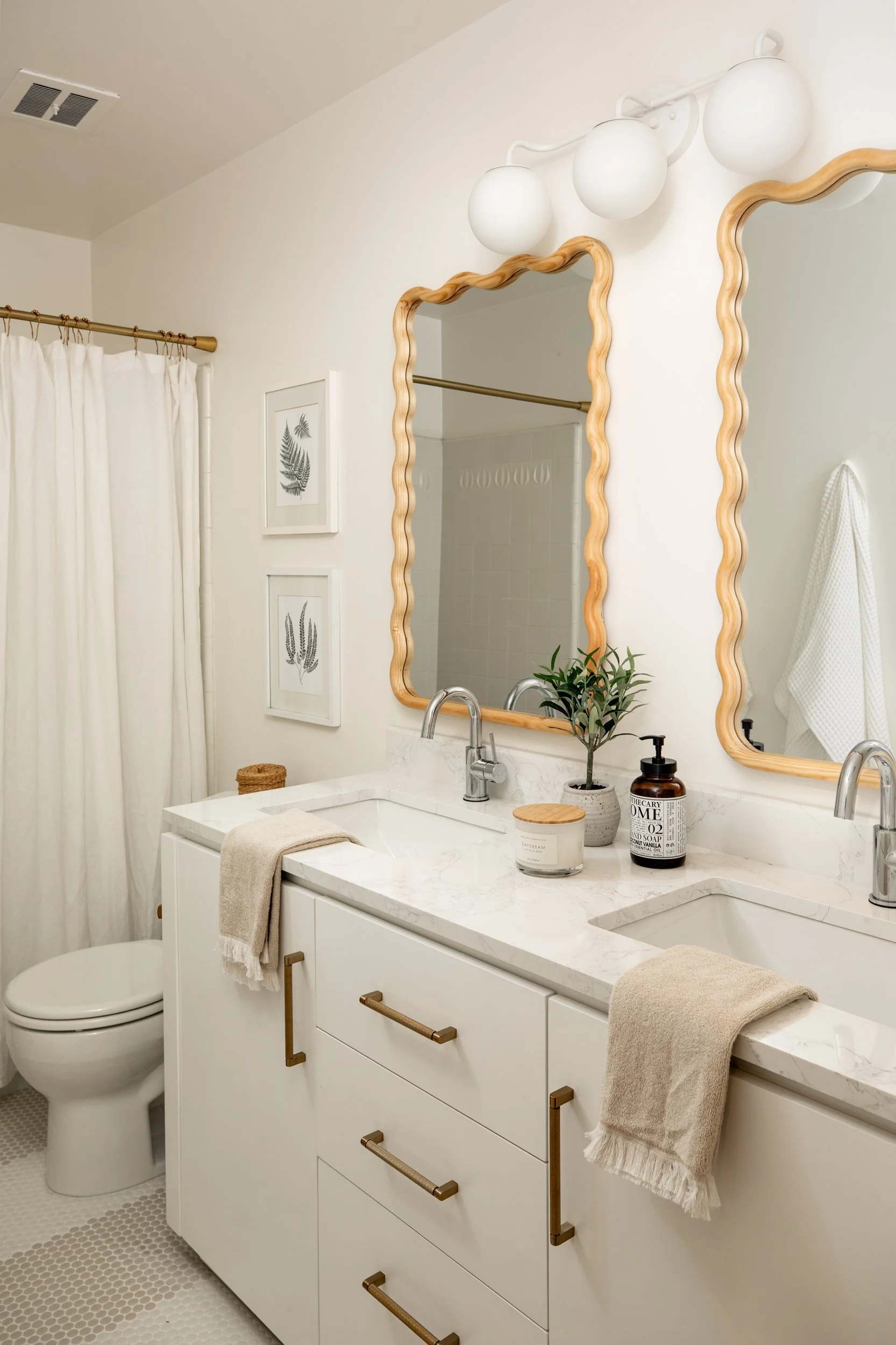 Bright bathroom with double mirror vanity, white marble countertop, potted plant, soap dispenser, candles, and towels, with framed botanical prints and a shower curtain.