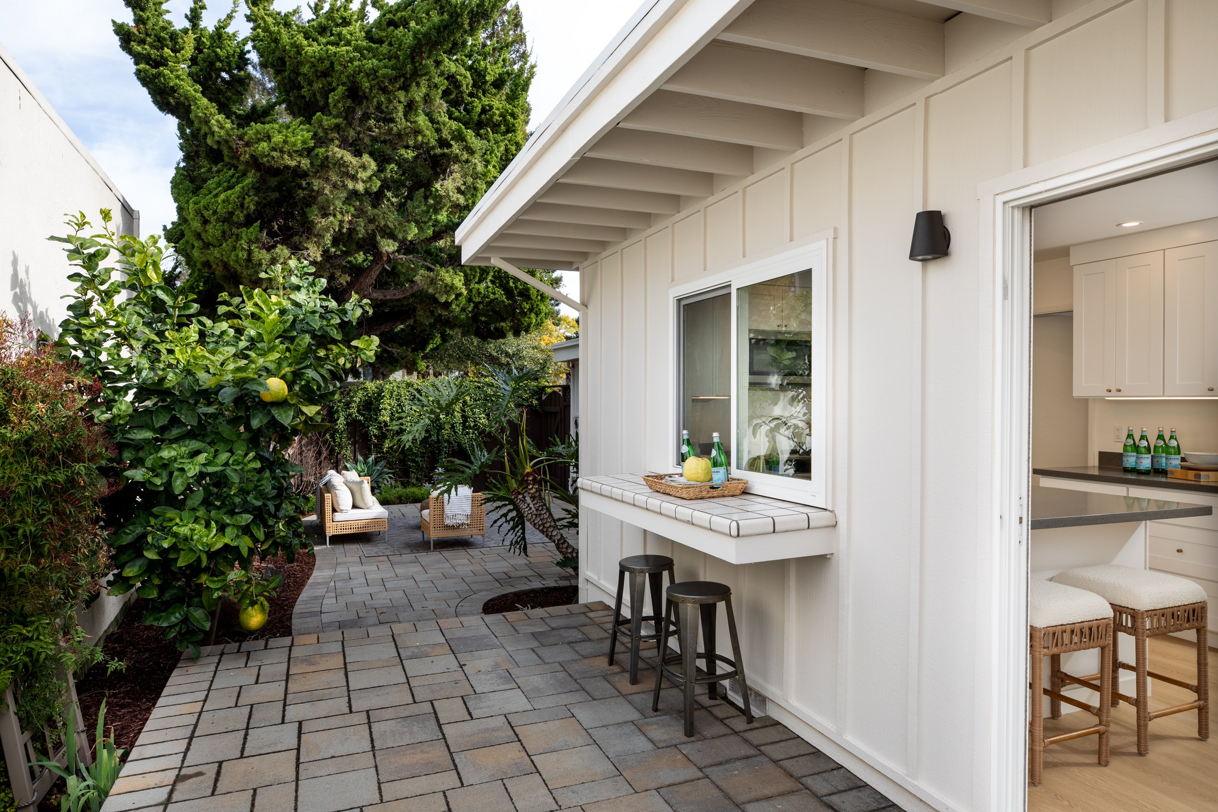 A cozy outdoor patio area with potted plants, a tree, and seating, connected to a white house with a window and an open door leading into a kitchen with bar stools.
