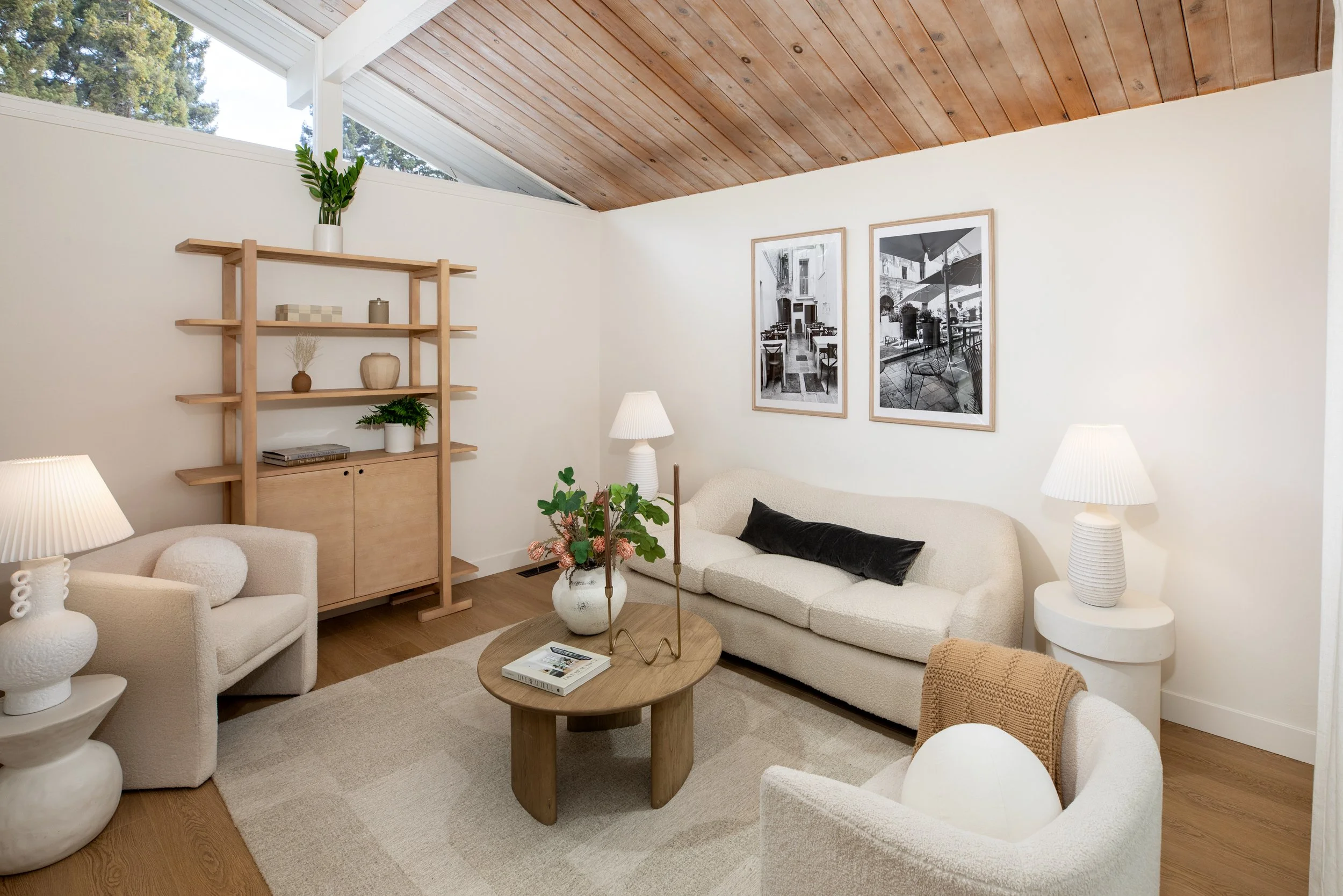 Living room with cream-colored sofa, armchairs, wooden coffee table, and lamps, featuring black and white wall art, wooden ceiling, and large window with trees outside.