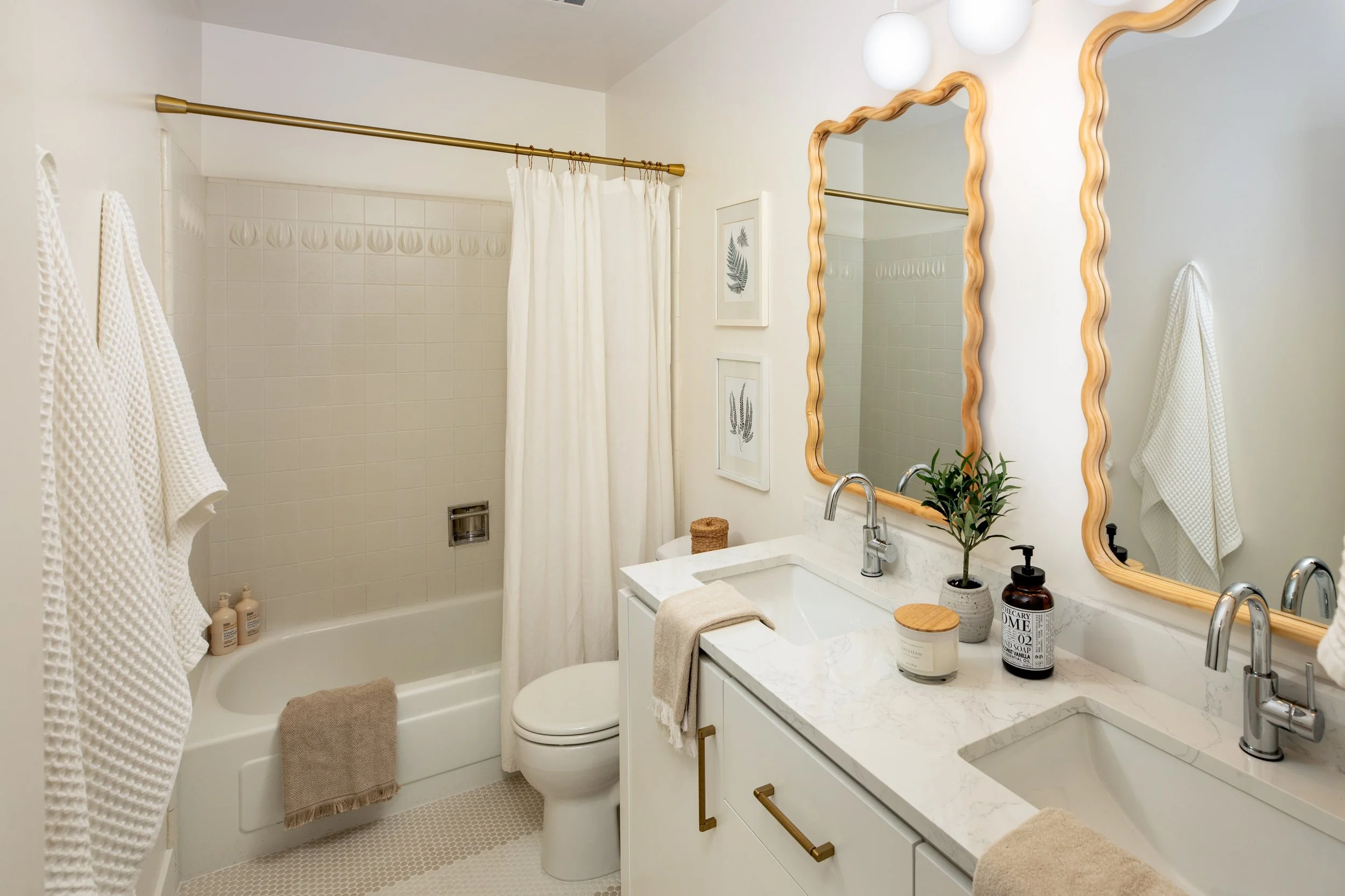A modern bathroom with a white marble double vanity, two large wavy-edged mirrors, a small potted plant, soap dispener, candles, and a beige towel. There are framed leaf prints on the wall, white towels hanging, and a bathtub with a white shower curt