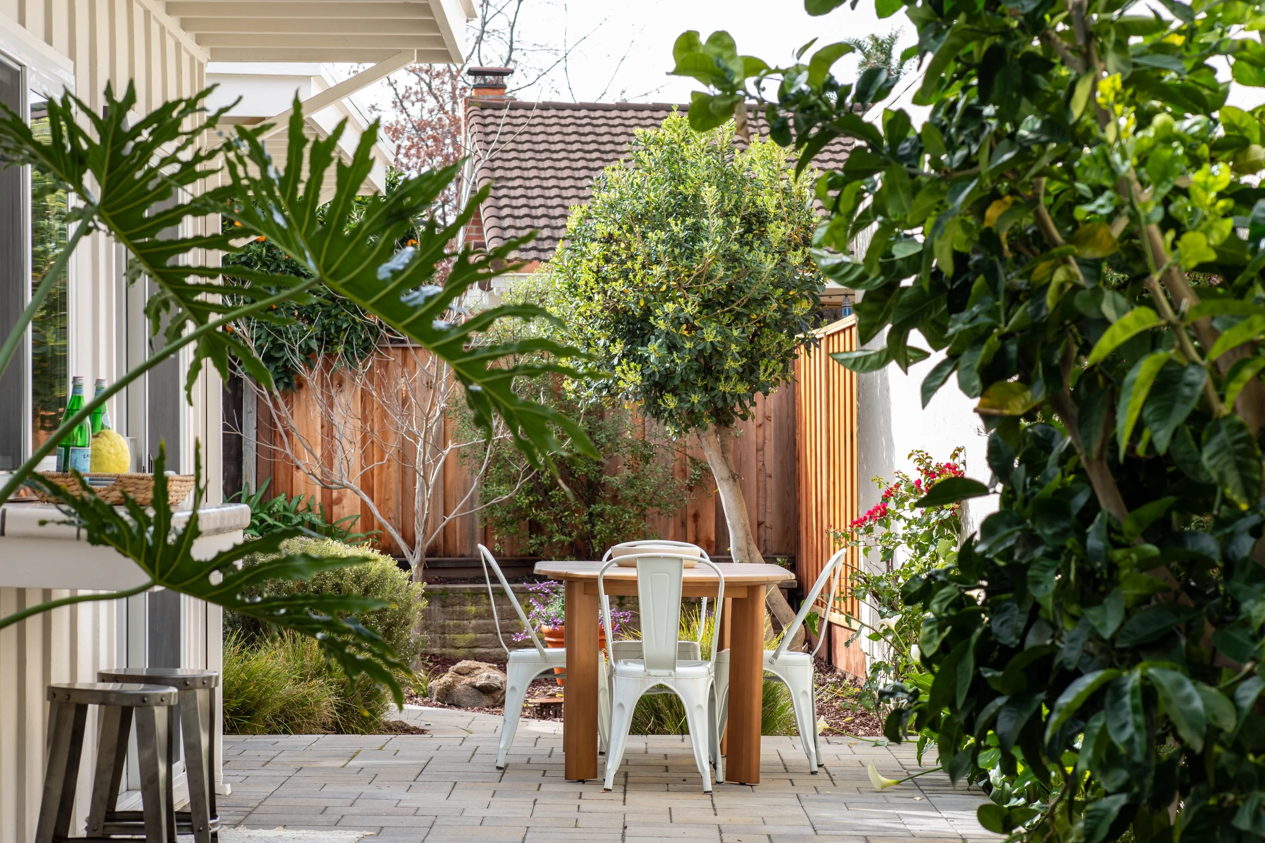 Outdoor patio area with a wooden table surrounded by white metal chairs, green trees and bushes, a wooden fence, a potted plant with pink flowers, and a house with beige siding and a brown roof in the background.