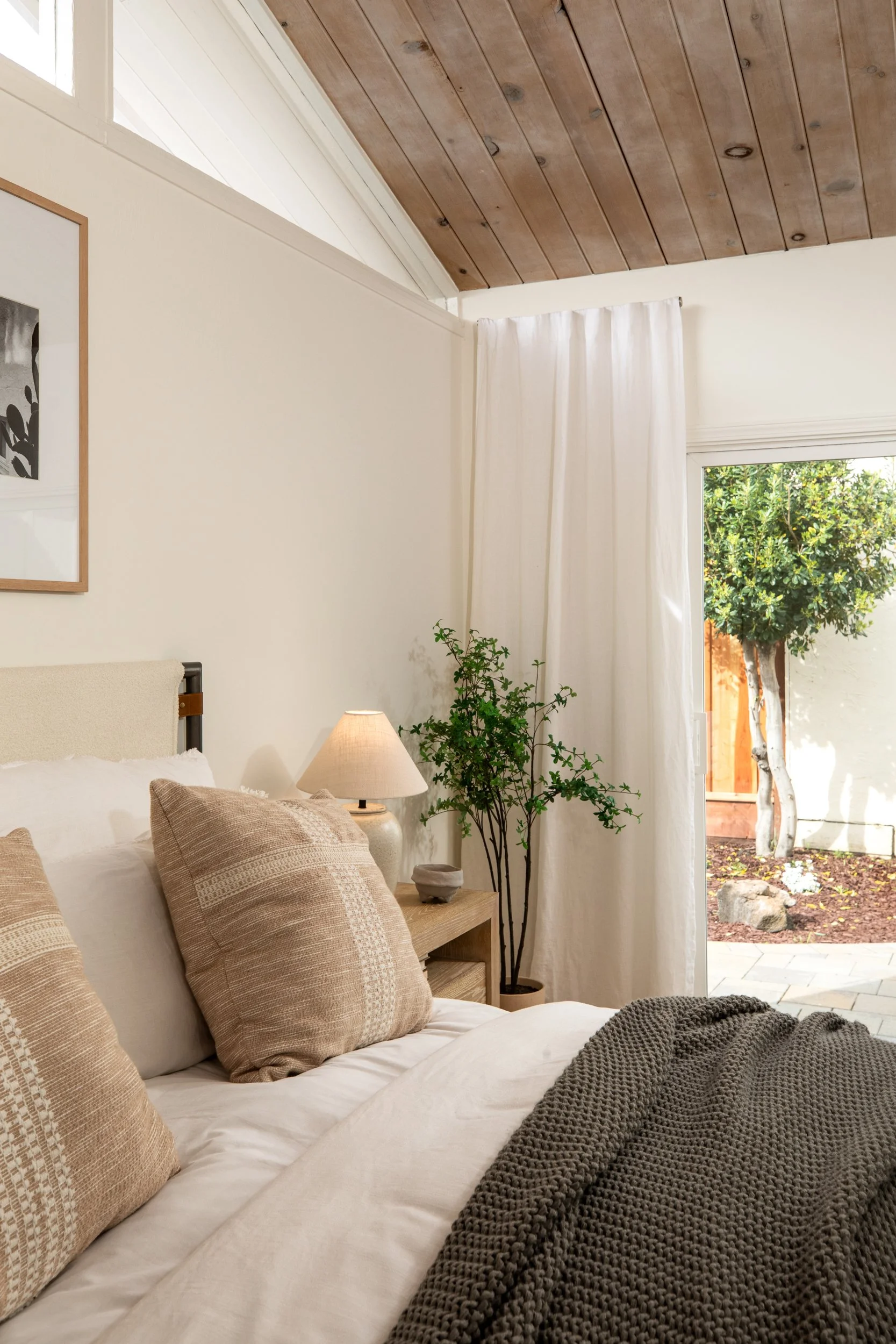 A cozy bedroom with beige pillows on a bed, a nightstand with a lamp and a potted plant, white curtains, and a sliding glass door leading outside to a garden with a tree.