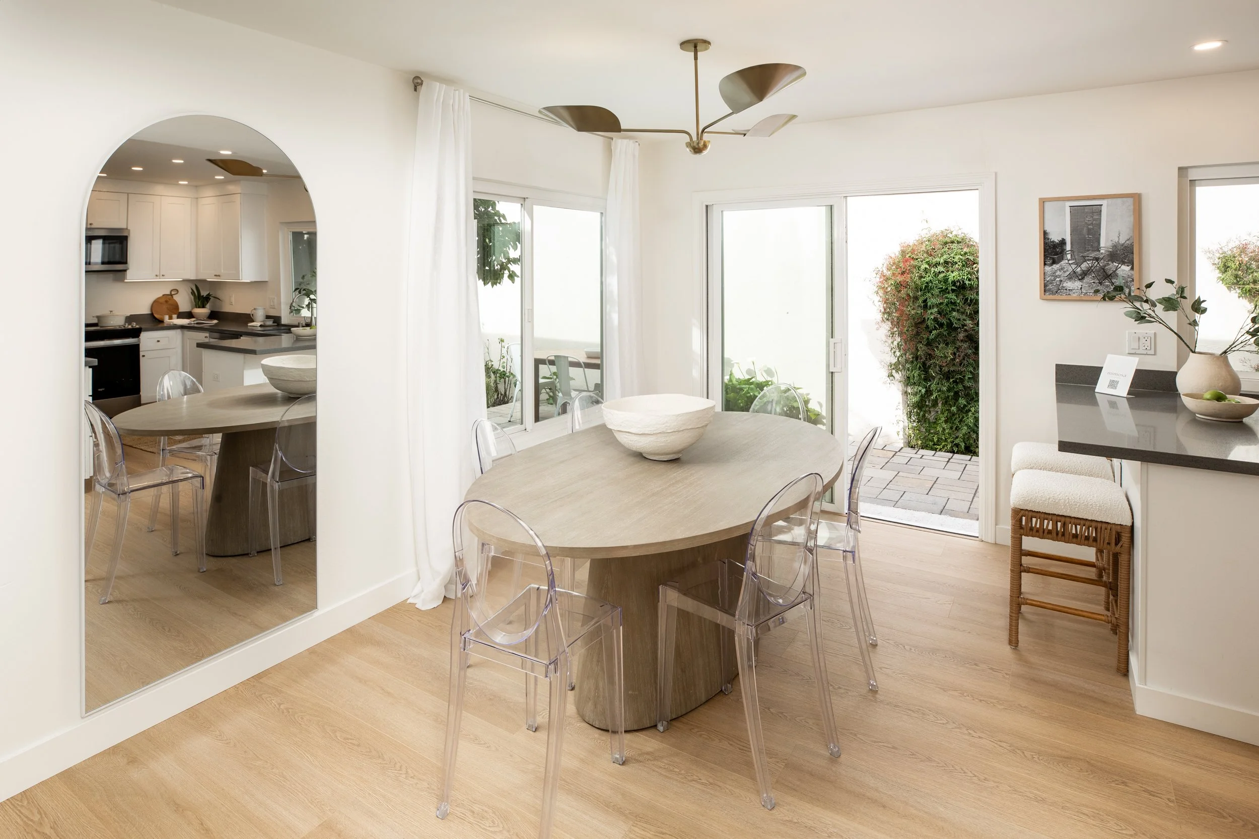 Bright dining room with round wooden table, six transparent chairs, sliding glass doors to patio, white curtains, and view of outdoor plants.