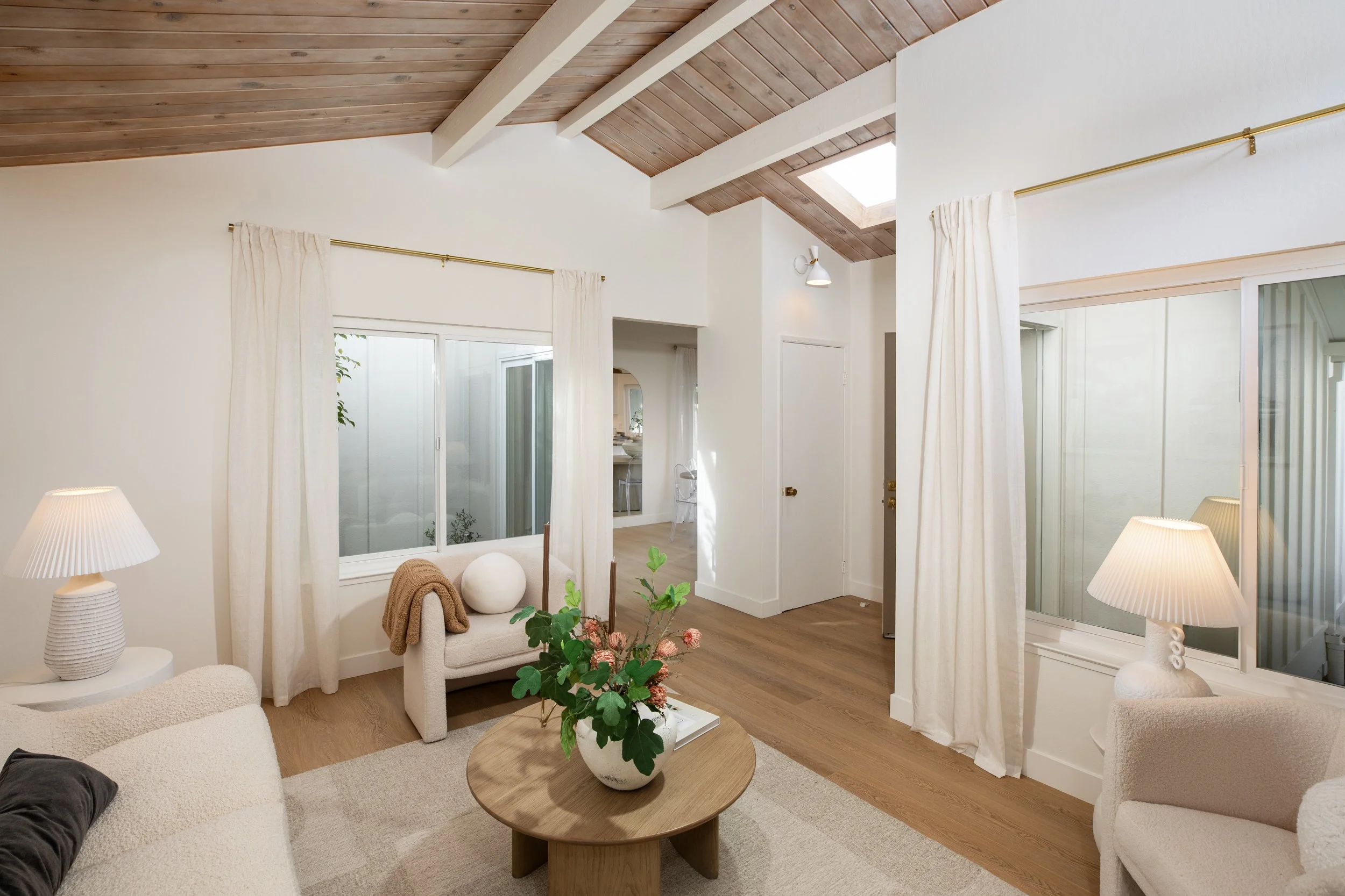 Living room with a wooden ceiling, white walls, beige furniture, curtains, and a round wooden coffee table with a flower arrangement.